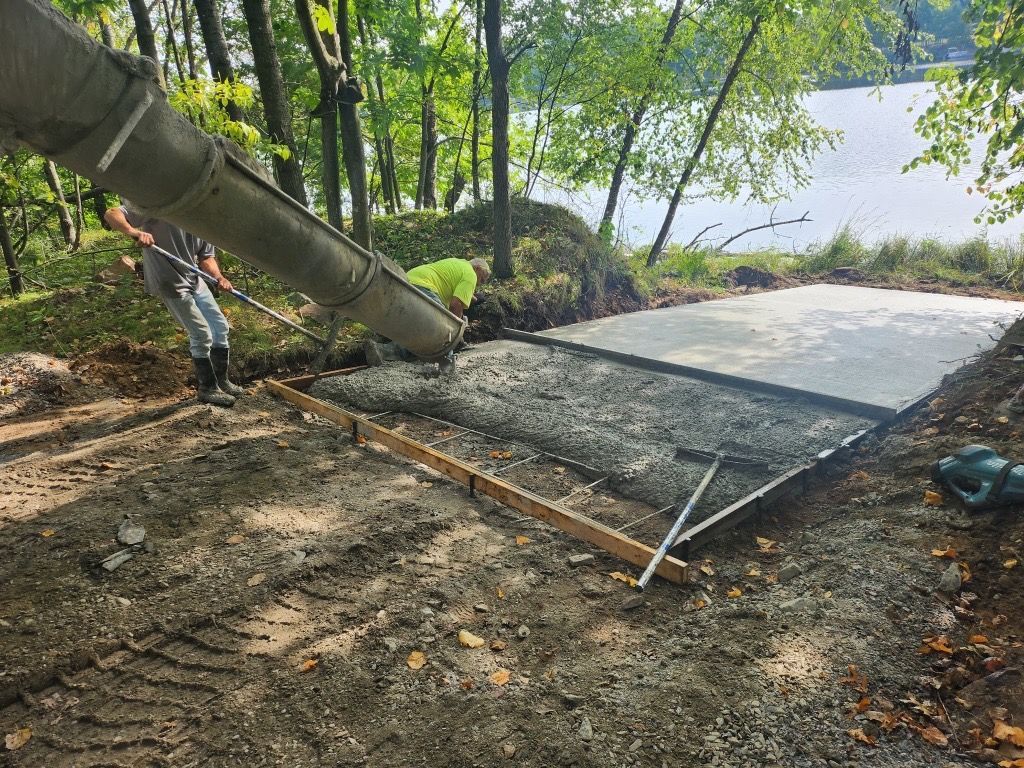 A metal rebar cage for a concrete pillar stands upright, embedded in a rectangular concrete footer at a construction site.