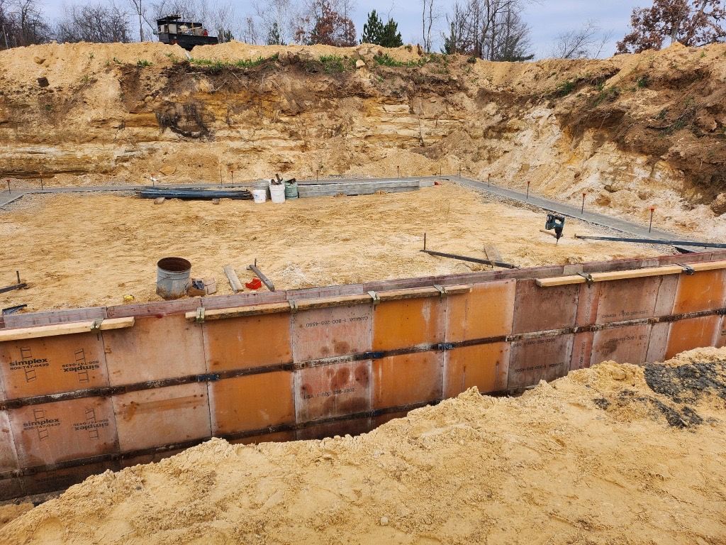 A yellow excavator rests on a construction site with dirt, rocks, and a worker in the background under a blue sky.