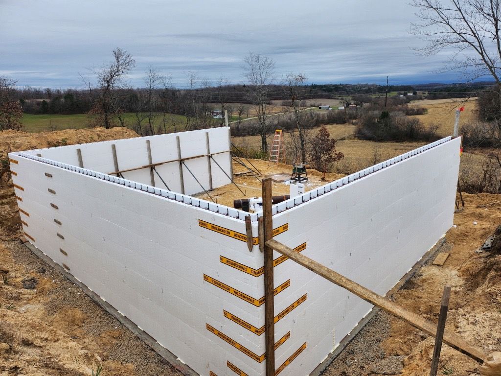 A yellow excavator sits beside a deep, rectangular trench revealing layers of red and tan soil on a construction site.