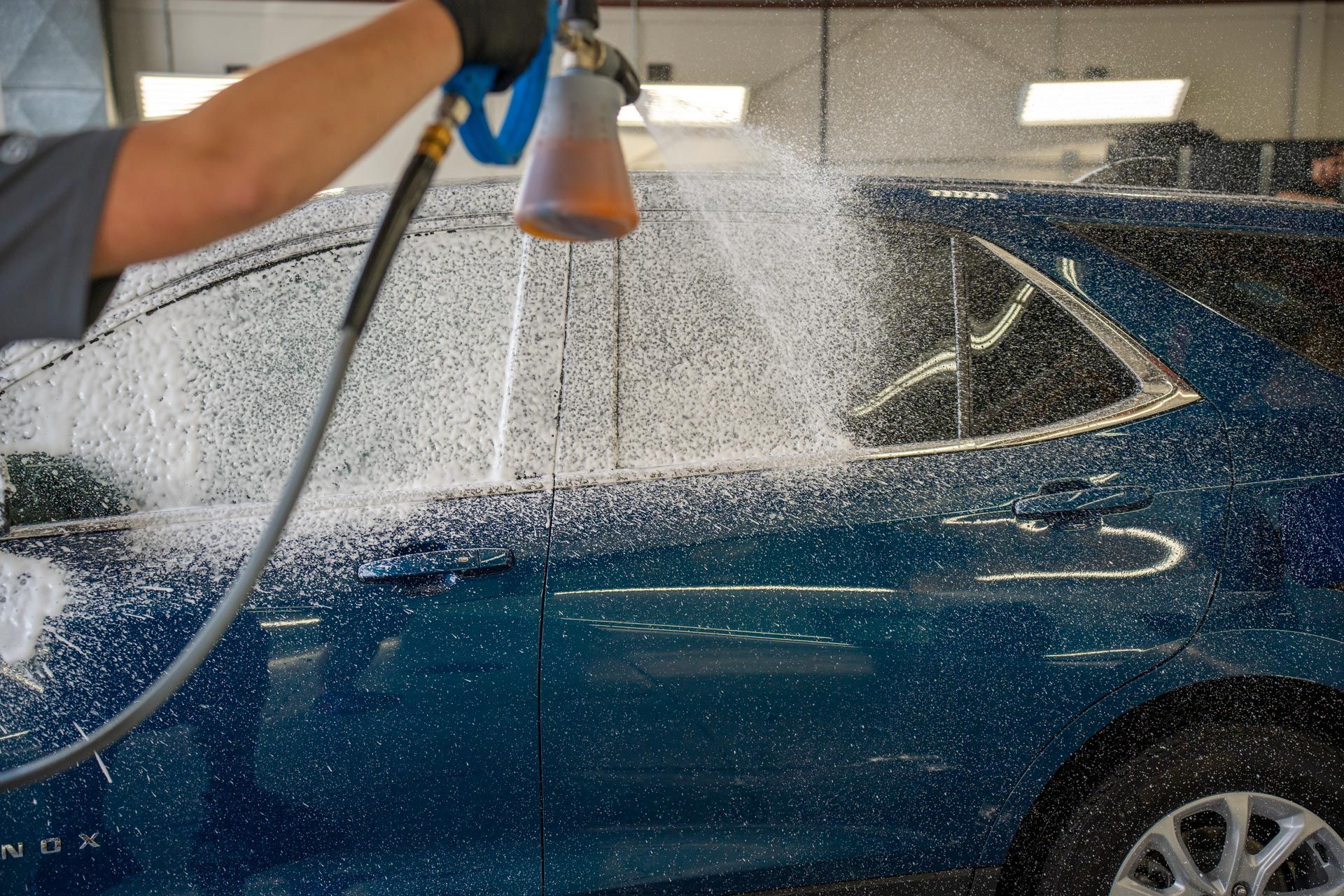 A person uses a foam cannon to spray white cleaning suds onto the side of a blue car in a garage.