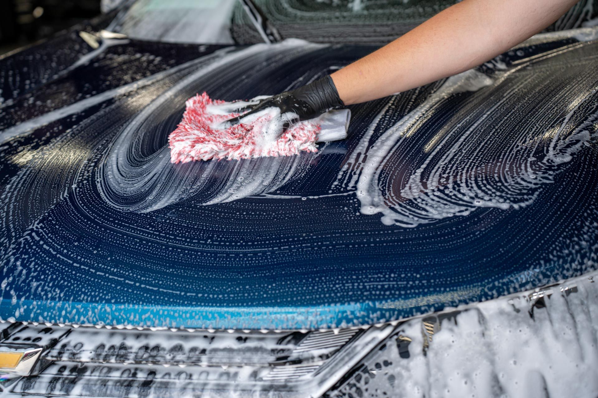 A hand in a black glove uses a red-and-white sponge to wash soap suds off the hood of a blue car.