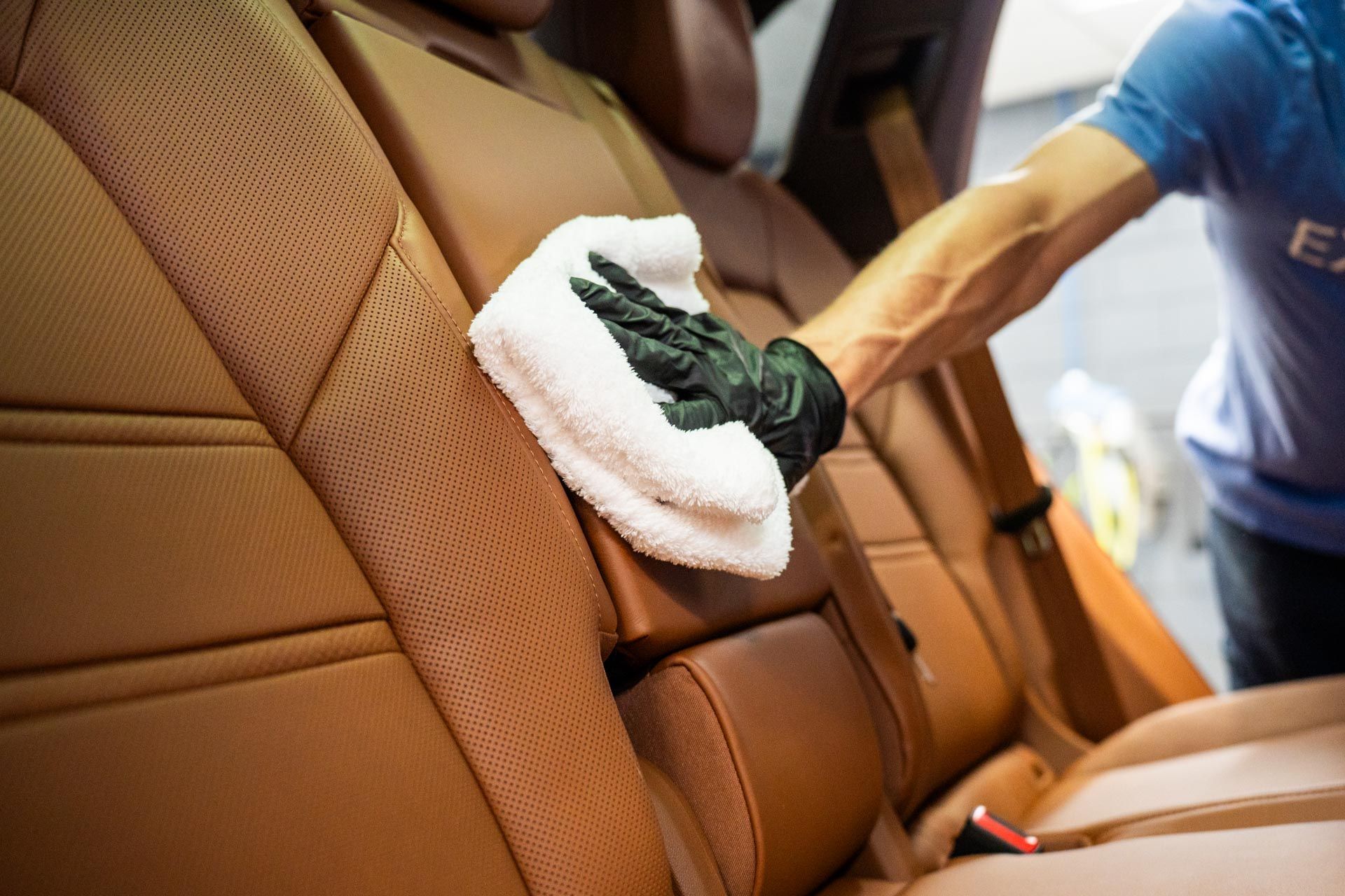 A person wearing a black glove uses a white cloth to clean the tan leather upholstery of a car seat.