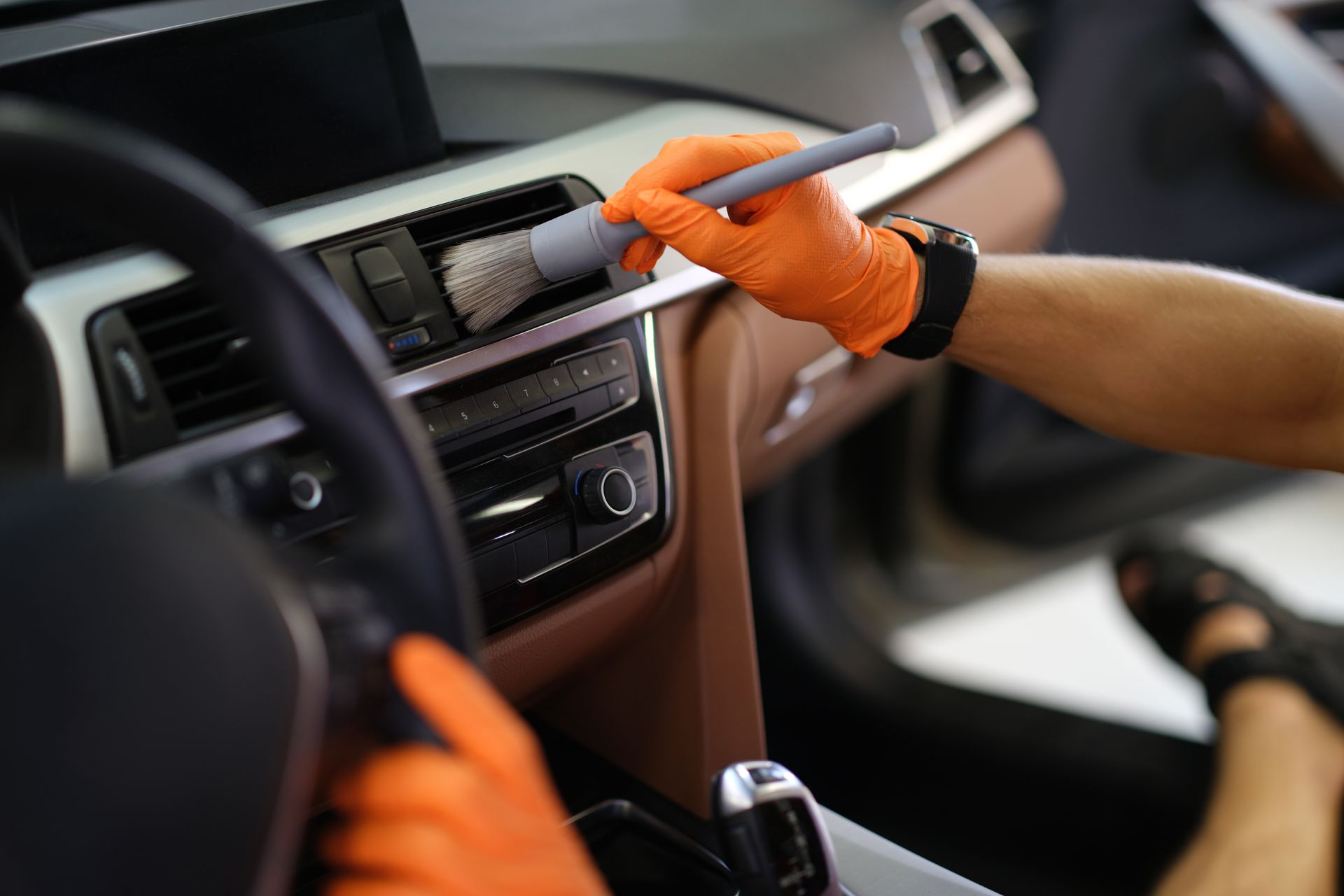 A hand in an orange glove uses a soft brush to clean the air vent of a car's dashboard.