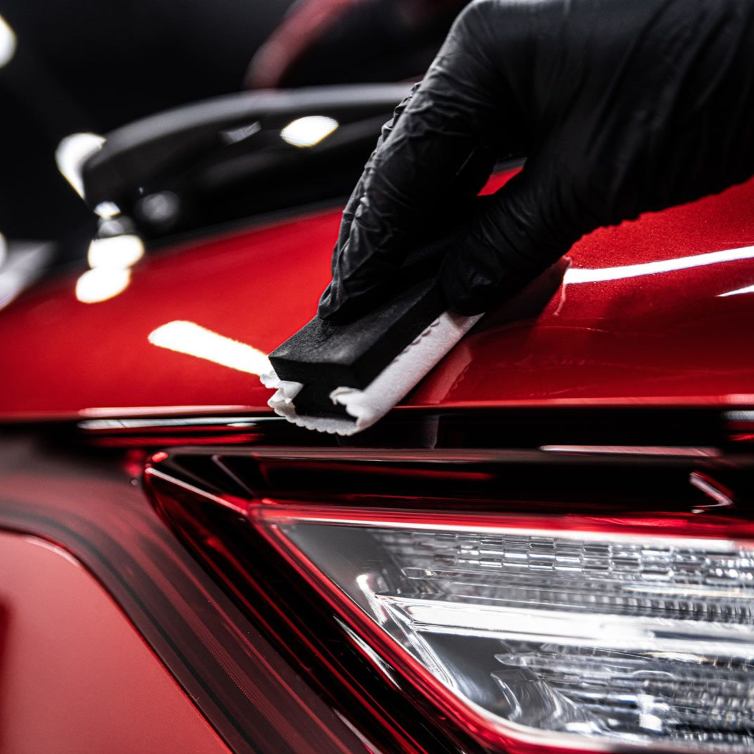 Technicians apply a clear protective film onto the hood of a black car in a garage with specialized overhead lighting.