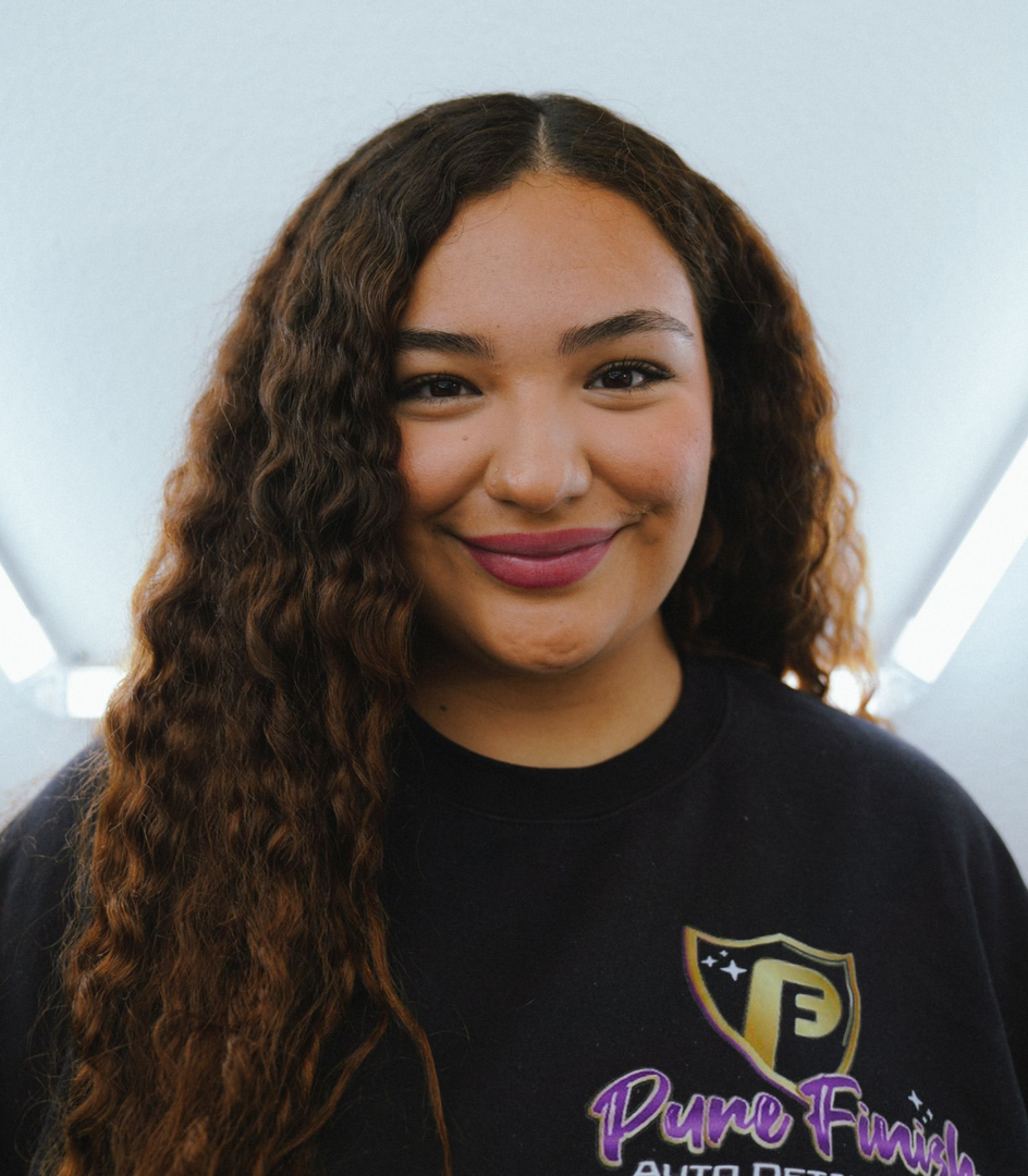 A person in a purple shirt uses a spray bottle to clean the soapy black rim and tire of a parked dark-colored vehicle.