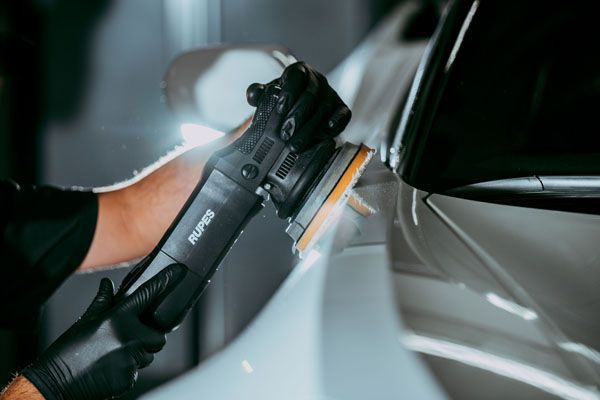 A person wearing black gloves uses a RUPES machine polisher on the light-colored fender of a car.