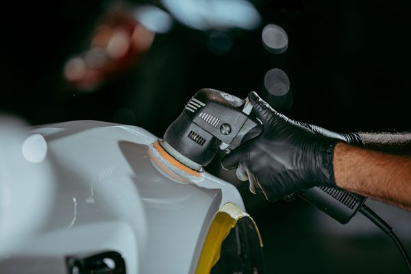 A gloved hand uses a power polisher on the white, curved surface of a vehicle in a dark garage.