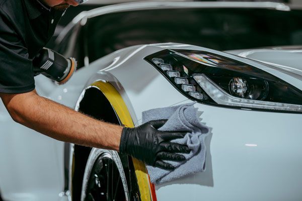 A person kneels to apply a protective film to the front bumper of a blue sports car in an auto shop.