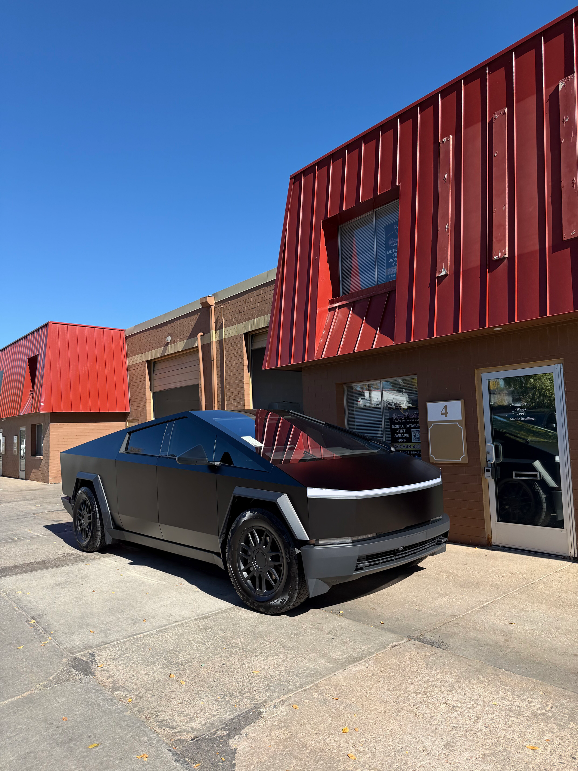 A matte black Tesla Cybertruck parked on a paved lot in front of a building with red metal siding.