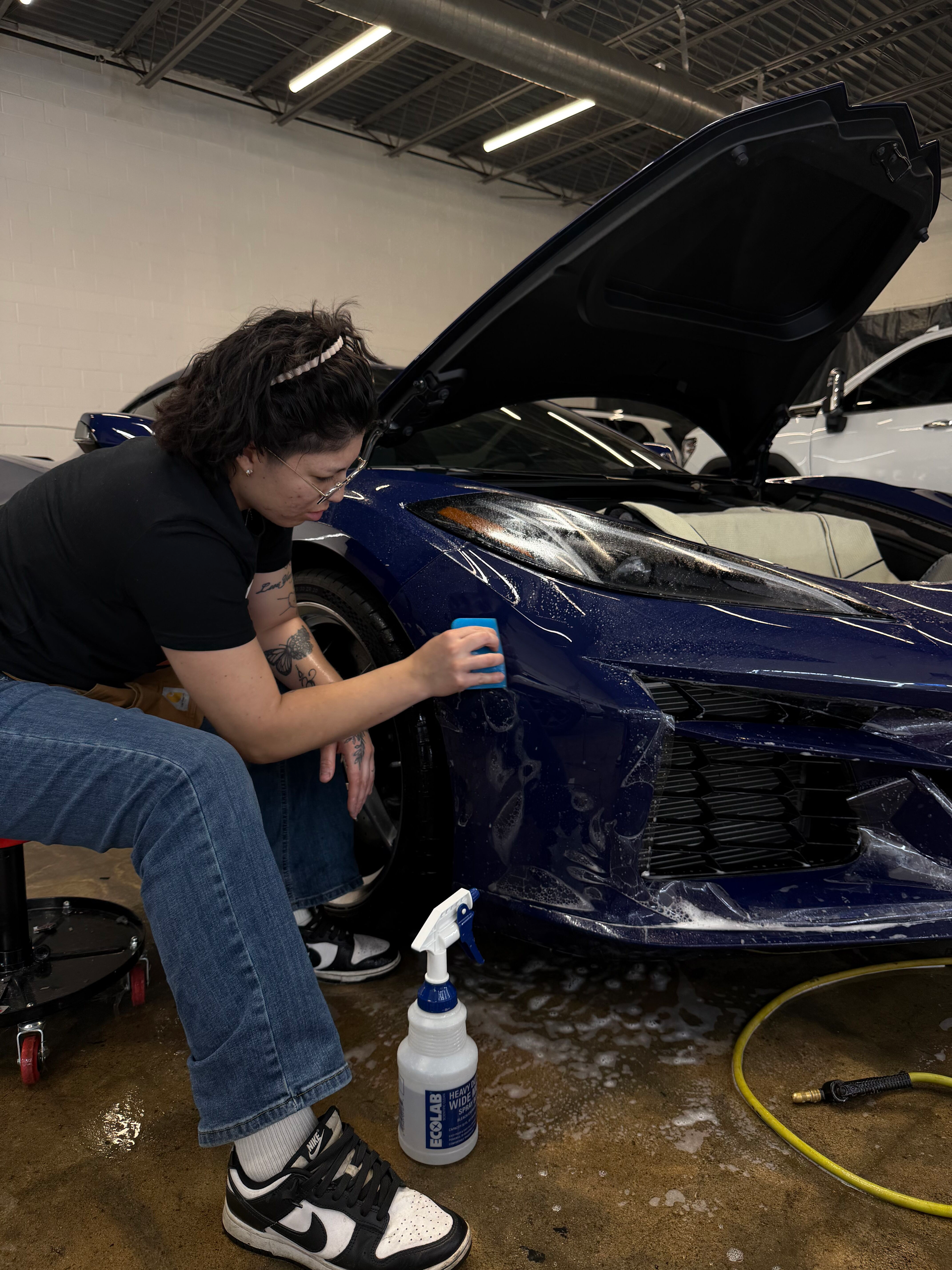 A person kneels to apply a protective film to the front bumper of a blue sports car in an auto shop.