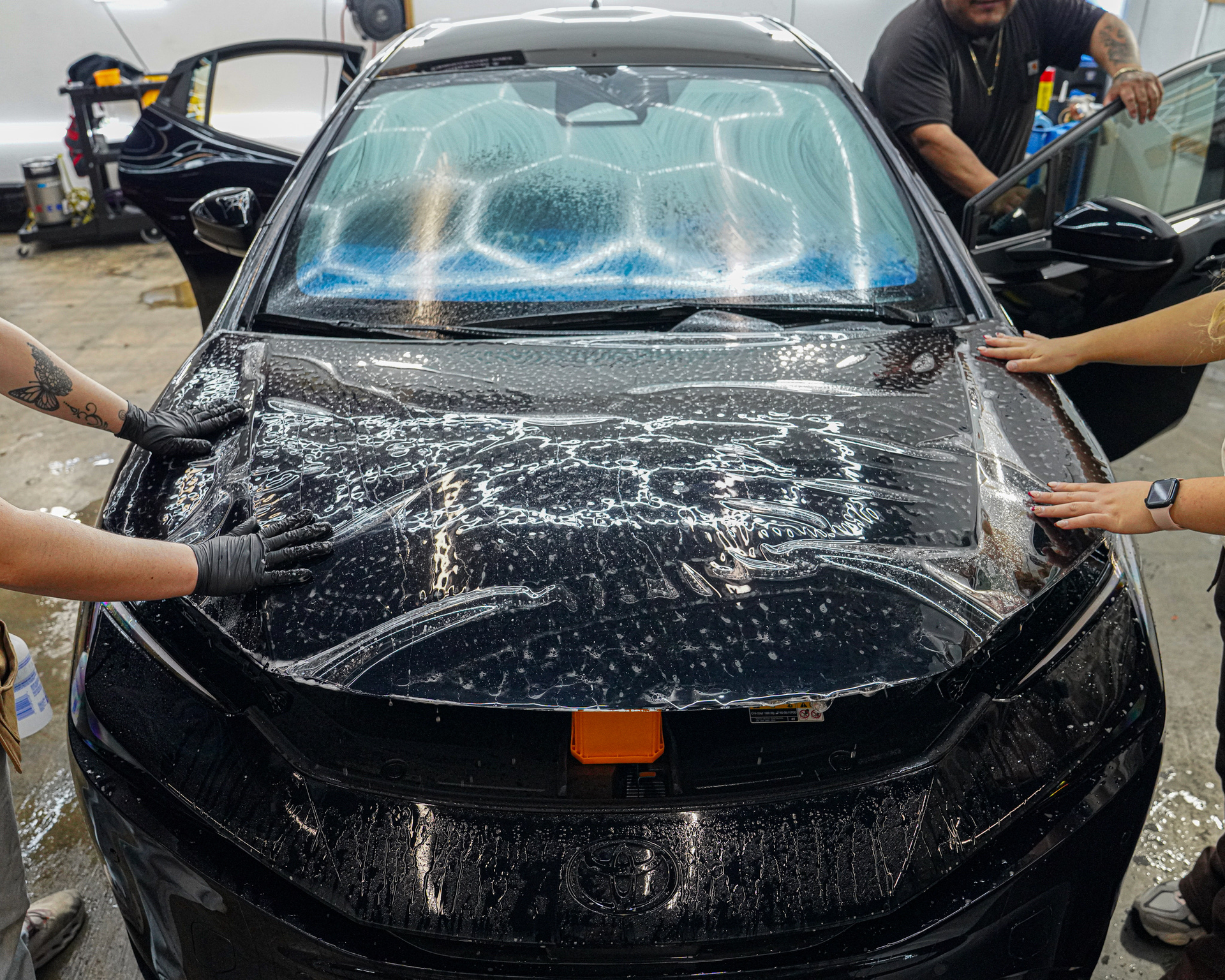 Technicians apply a clear protective film onto the hood of a black car in a garage with specialized overhead lighting.