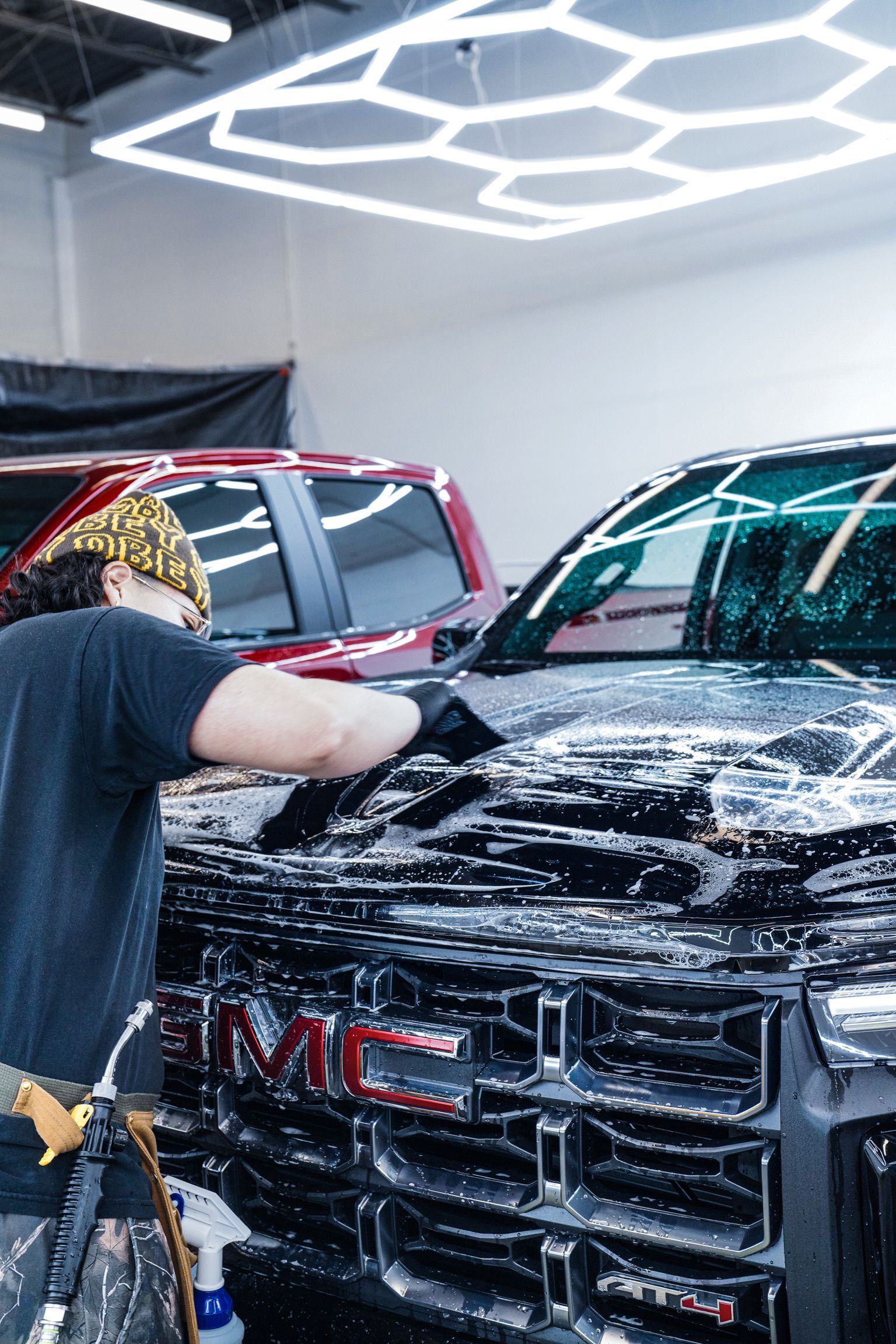 A worker in a shop washes the front of a black GMC truck covered in soap suds, with a red vehicle parked behind it.