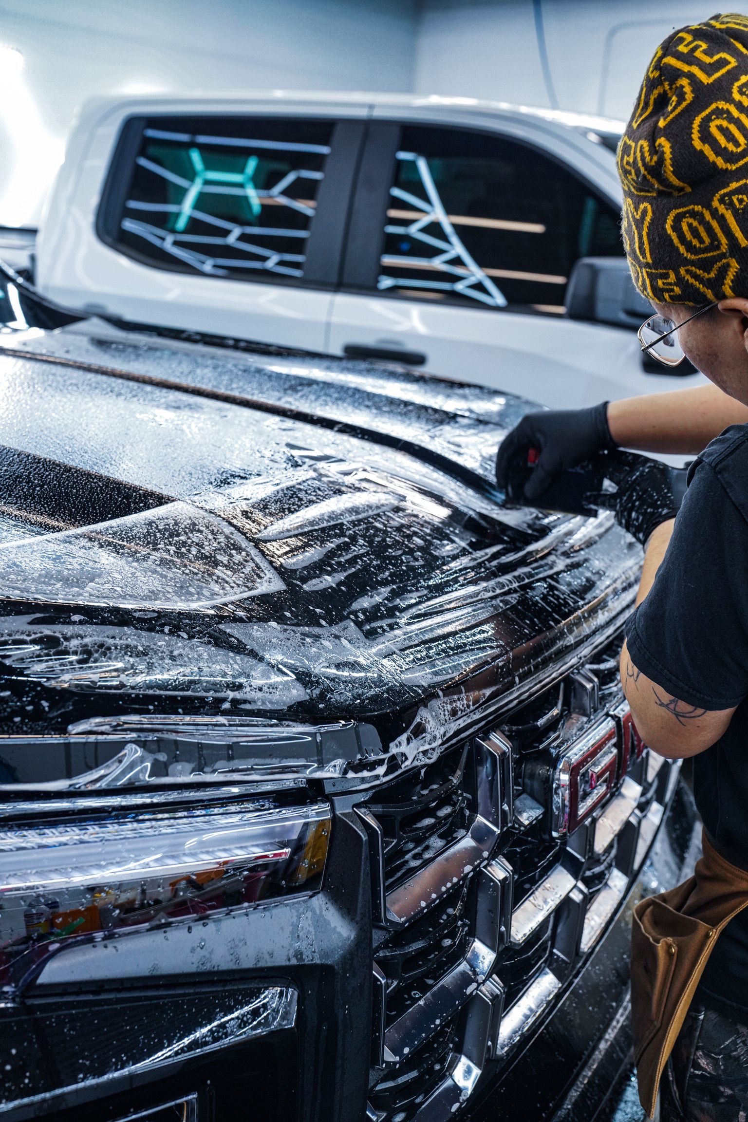 A person in a patterned beanie and black gloves applying a soapy cleaning film to the hood of a black vehicle.