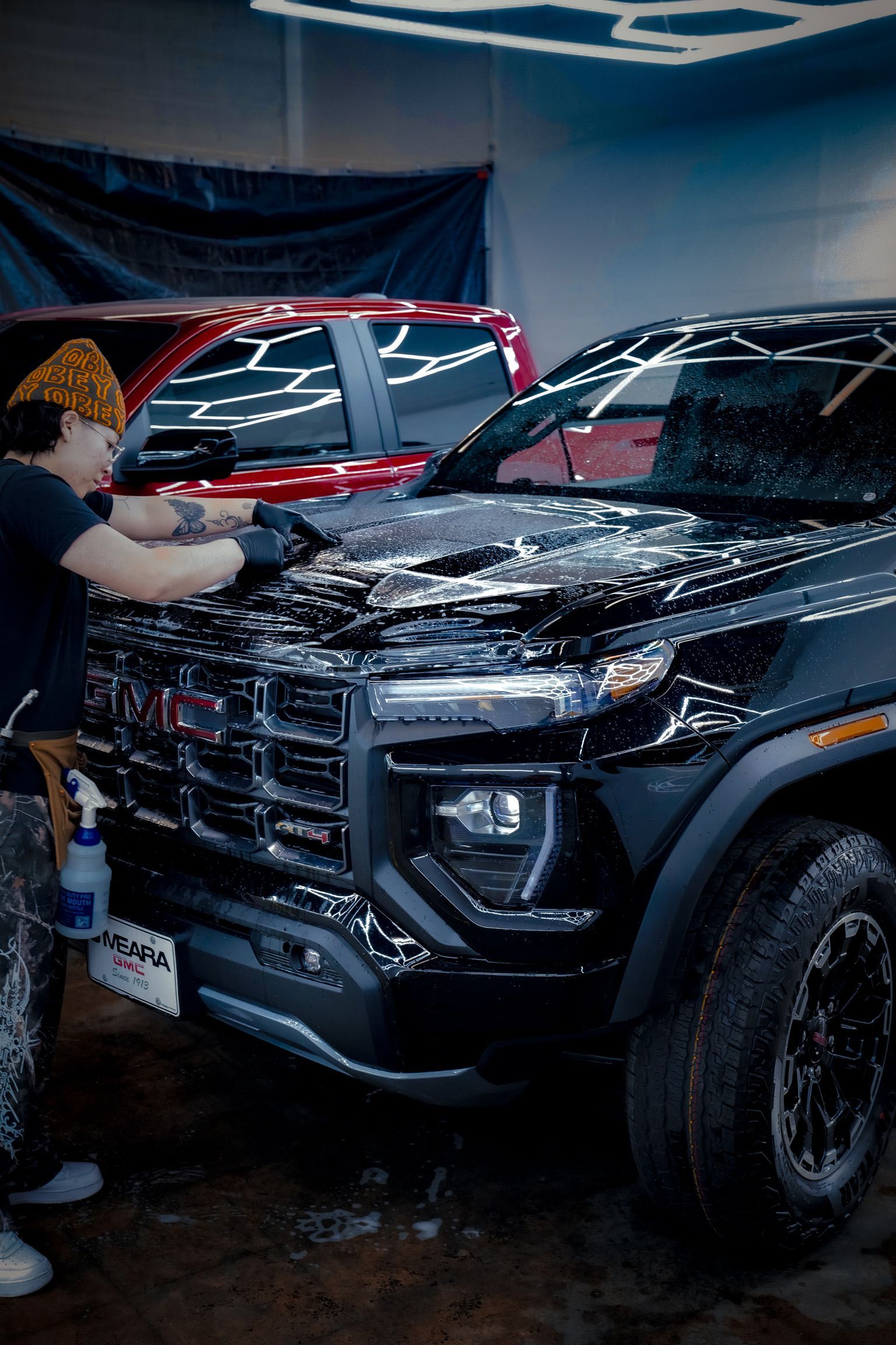 A person in a garage applying a protective film to the hood of a black GMC pickup truck, with a red truck visible nearby.
