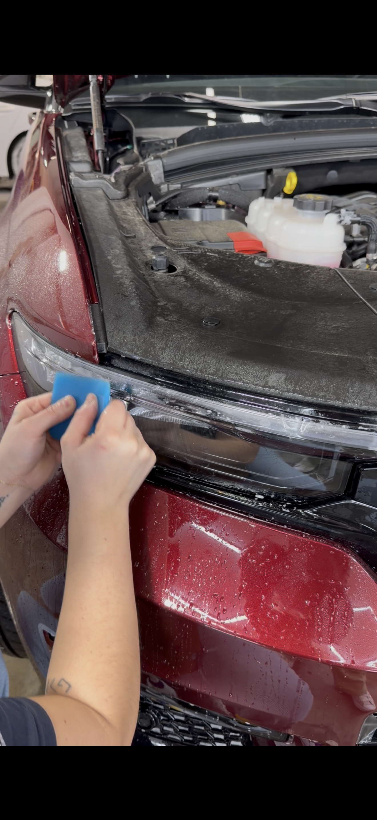 A hand uses a blue squeegee to apply a clear paint protection film to the hood of a gray car in a workshop.