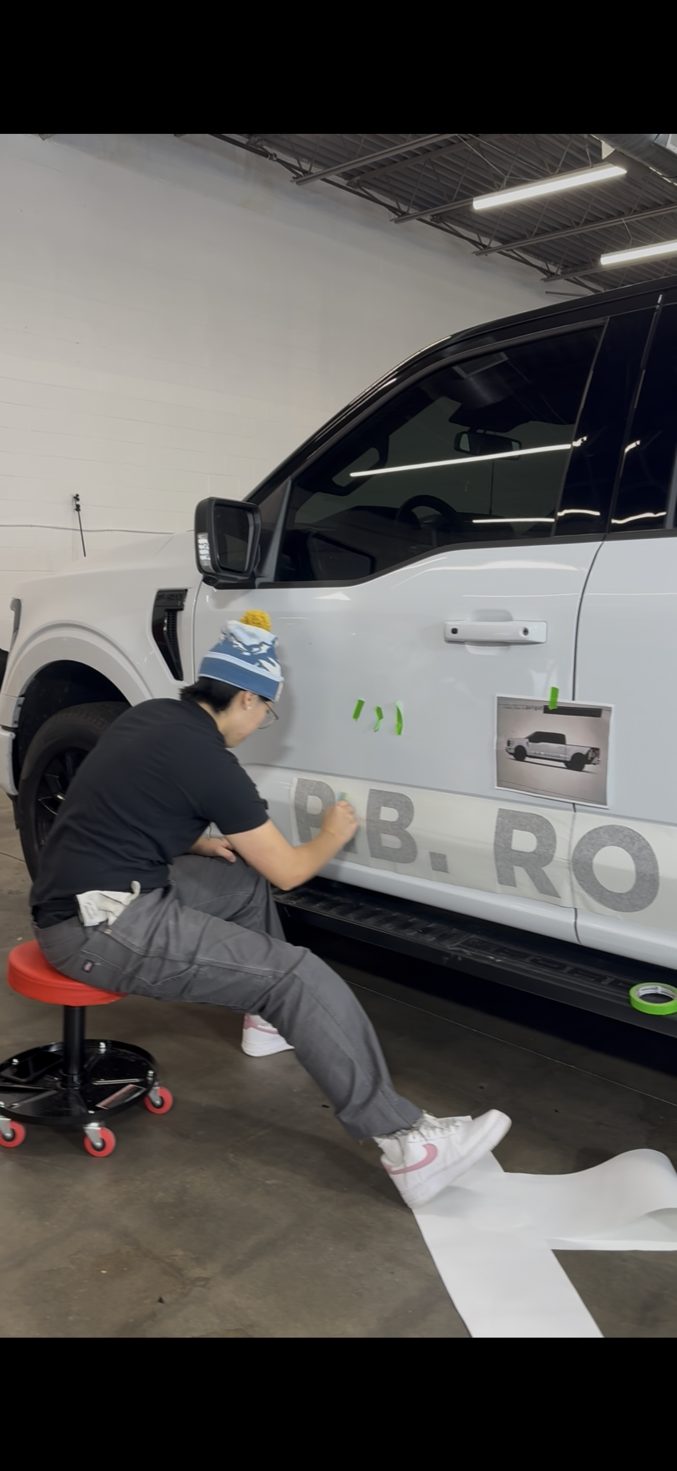 A person sits on a rolling stool, applying a large decal to the side of a white truck in a workshop.