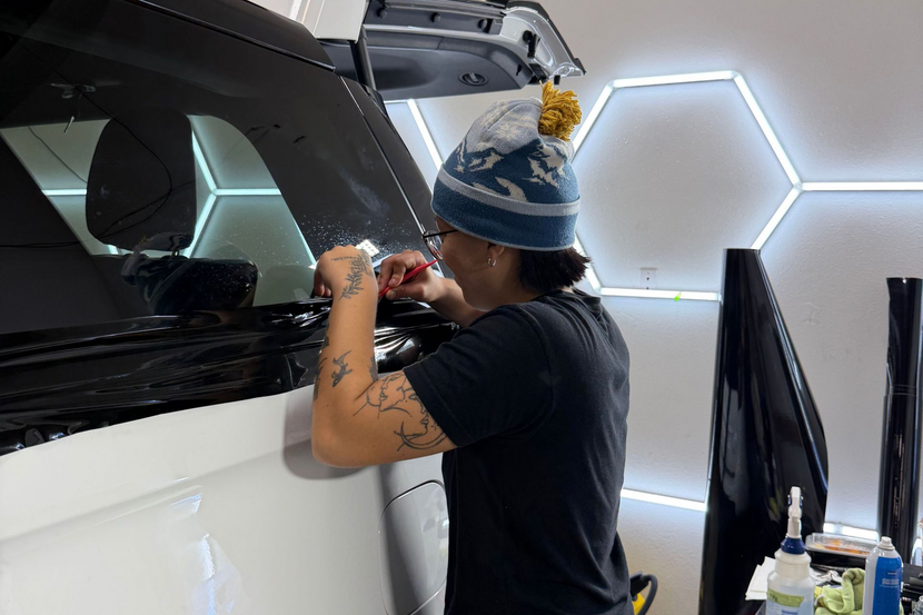 A person wearing a beanie carefully applies window tint film to the rear passenger window of a white vehicle.