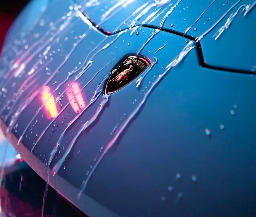 Technicians apply a clear protective film onto the hood of a black car in a garage with specialized overhead lighting.