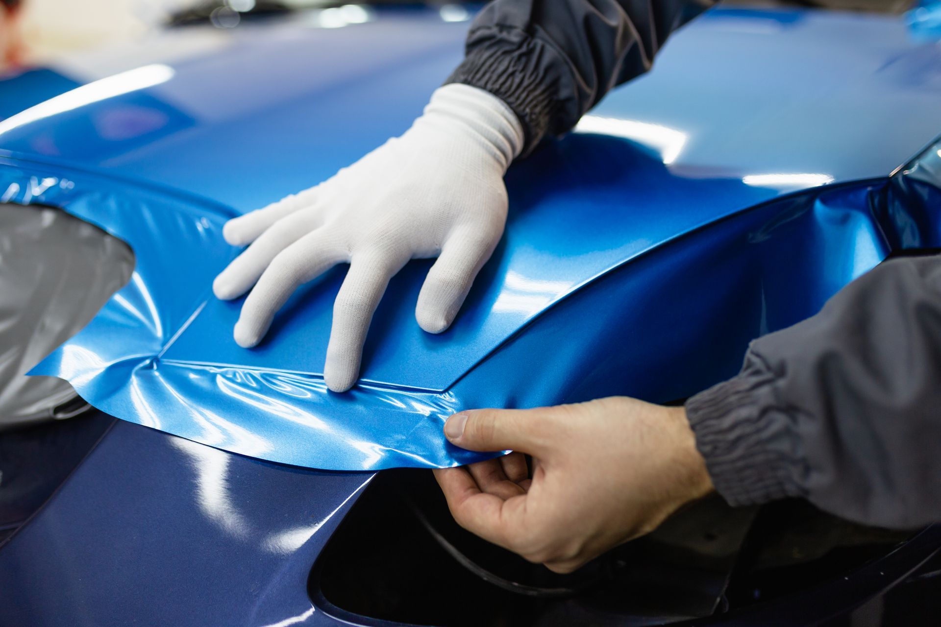 A person wearing white gloves applies a glossy blue vinyl wrap to a car hood.
