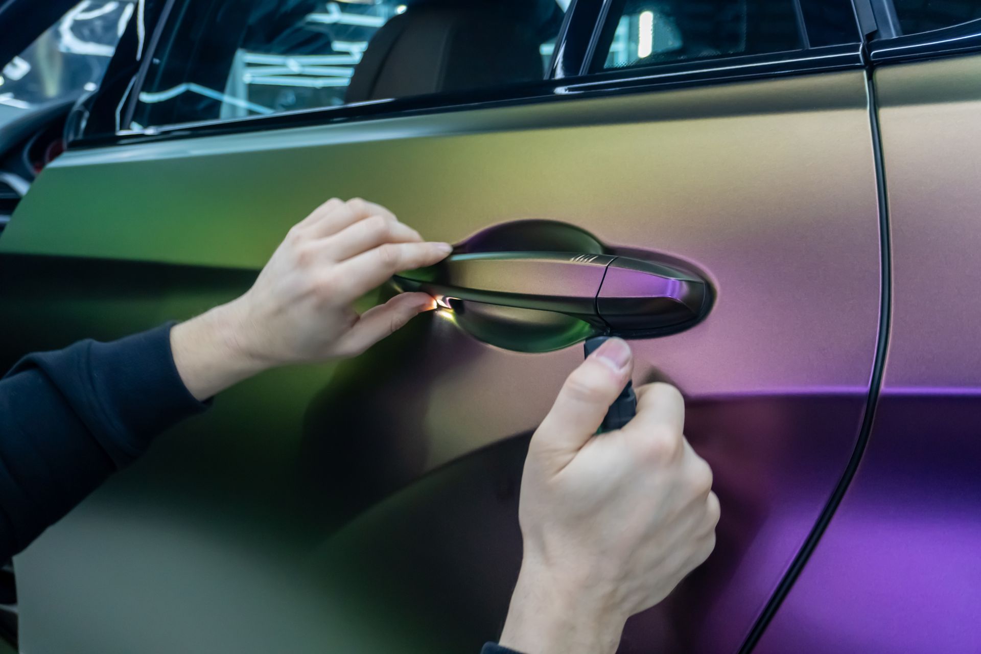 A person kneels to apply a protective film to the front bumper of a blue sports car in an auto shop.