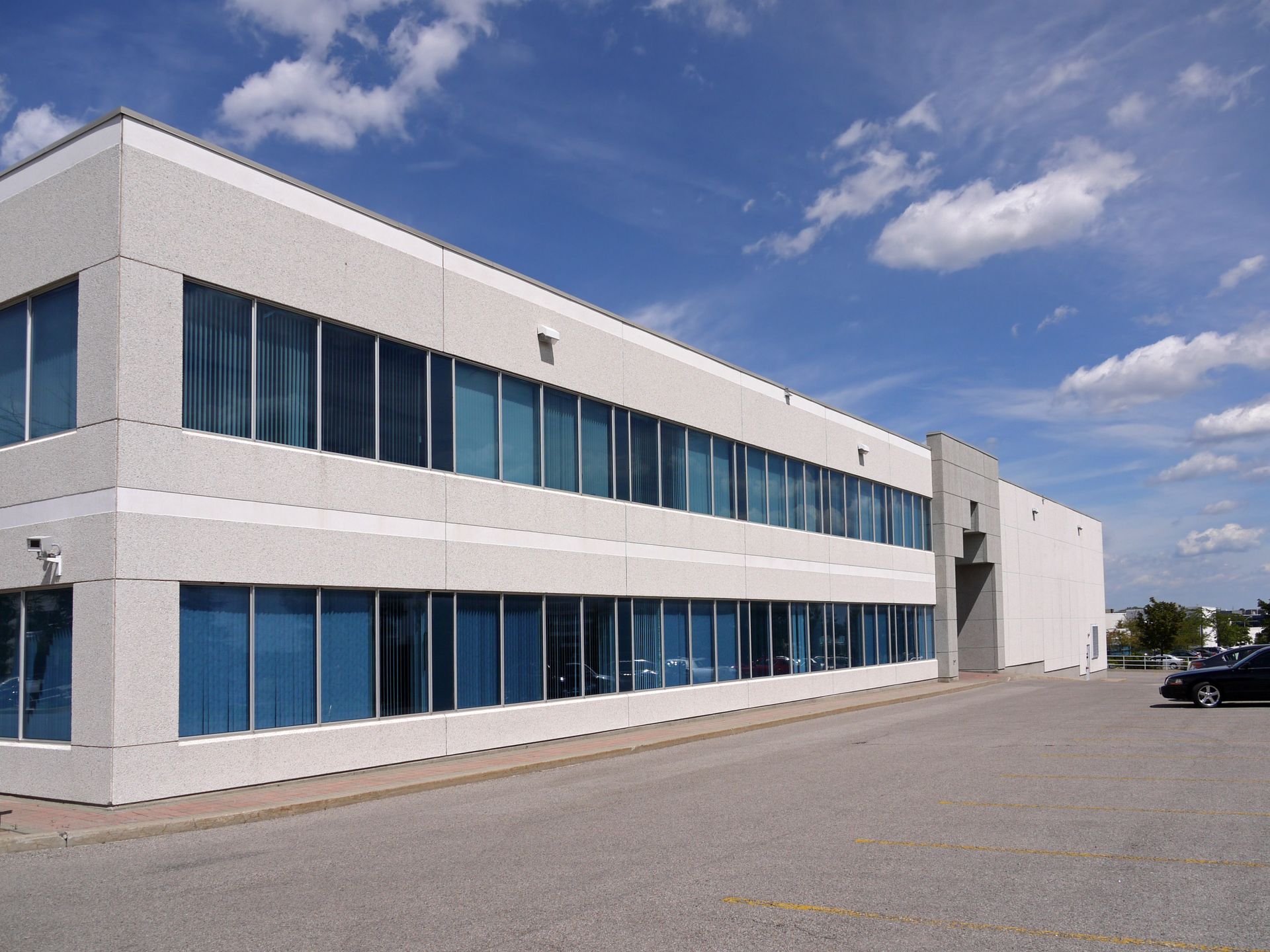 A two-story, rectangular office building with light grey concrete walls and rows of blue windows under a cloudy blue sky.