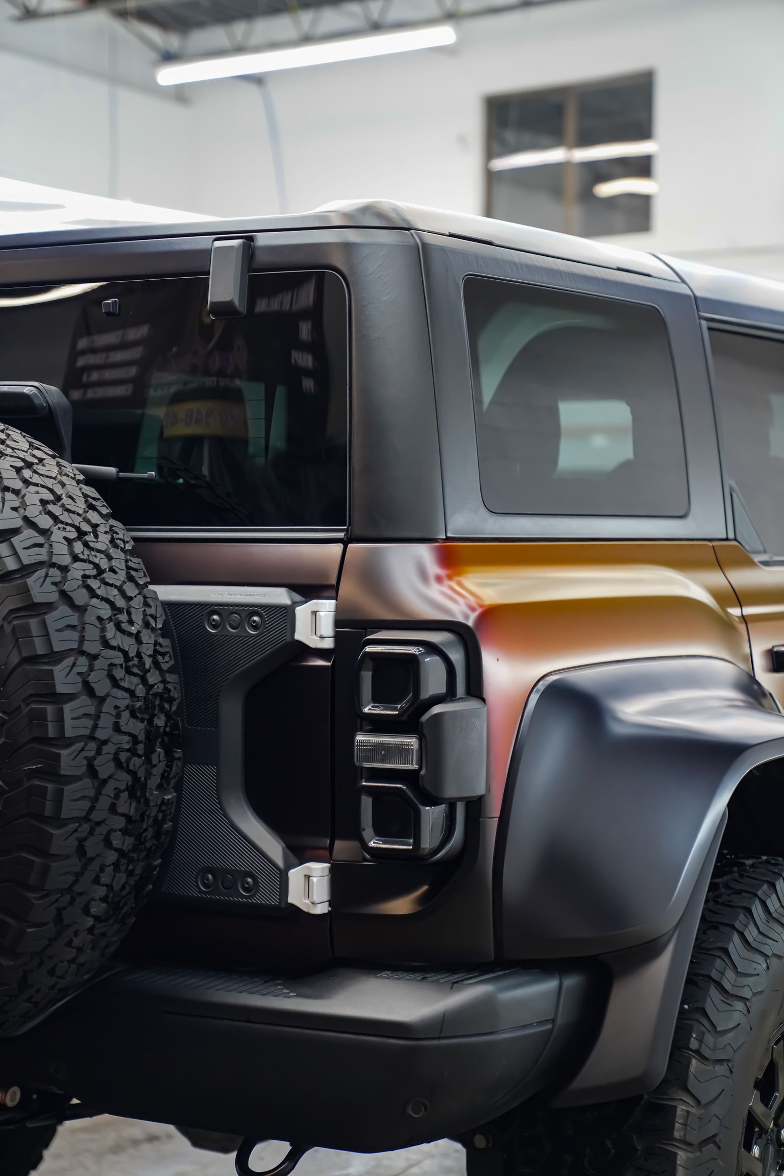 A person kneels to apply a protective film to the front bumper of a blue sports car in an auto shop.