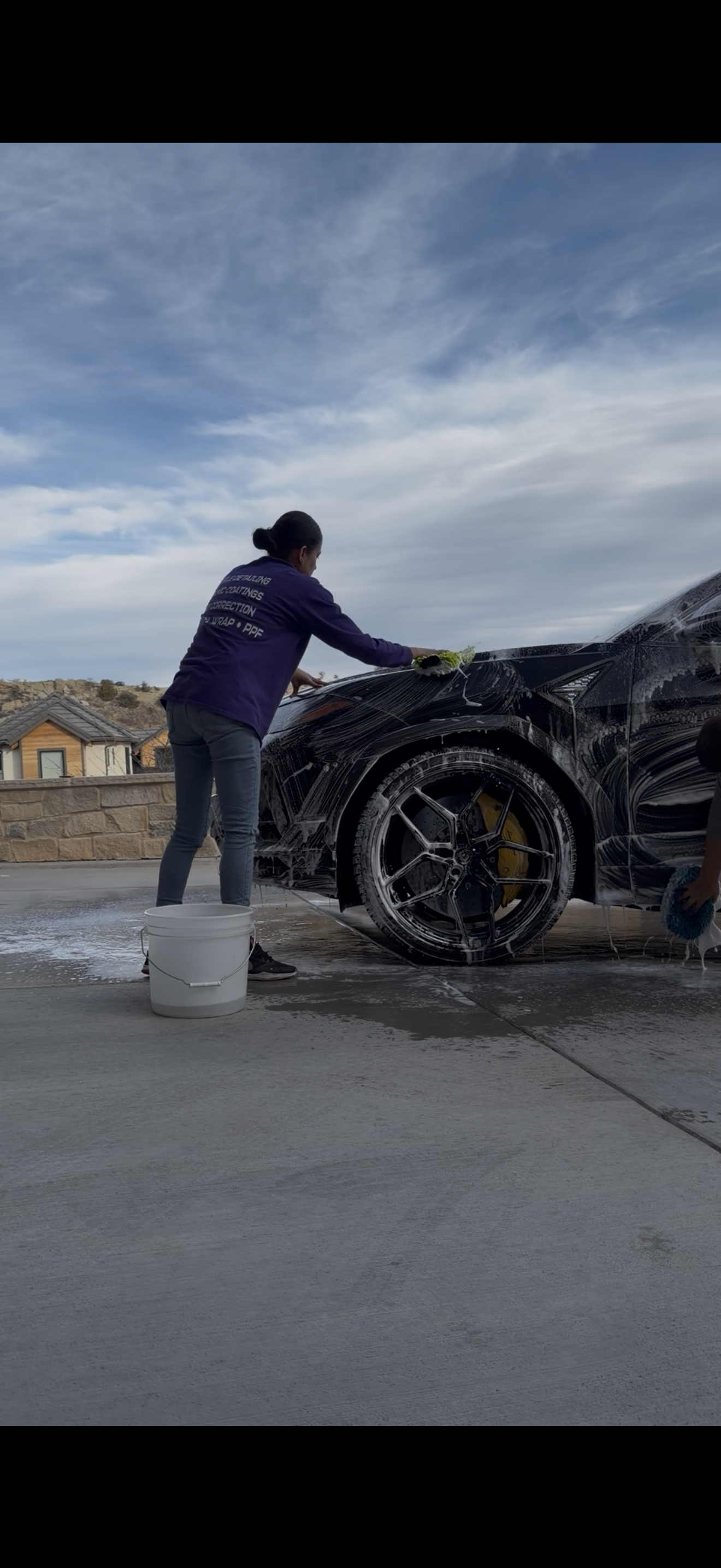 A person wearing a purple hoodie washing a soap-covered car on a driveway against a cloudy sky.