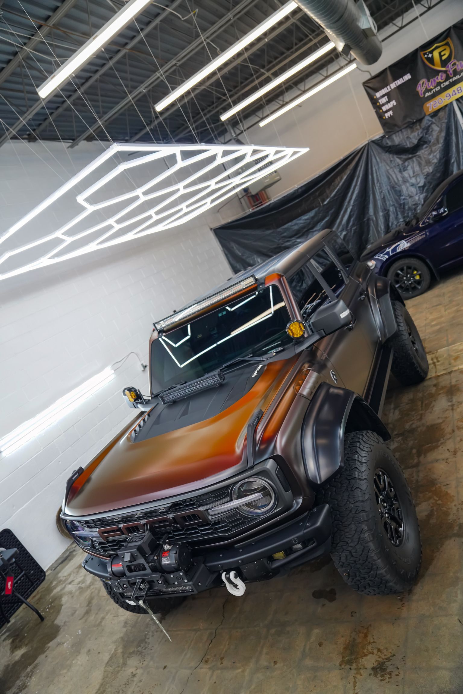 A modified Ford Bronco with a two-tone copper and black paint job parked in a workshop under hexagonal ceiling lights.