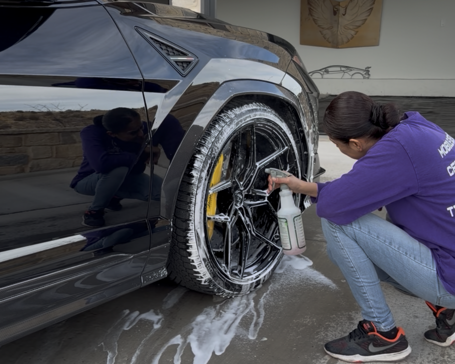 A person in a purple shirt uses a spray bottle to clean the soapy black rim and tire of a parked dark-colored vehicle.