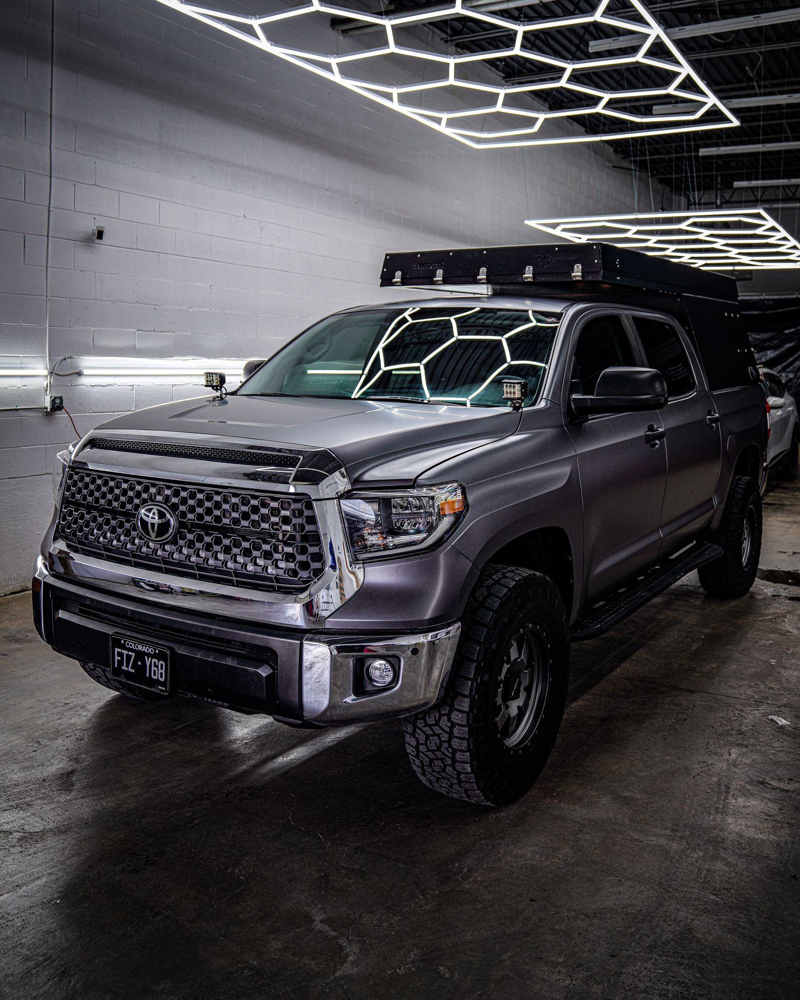 A dark gray Toyota Tundra pickup truck with an off-road roof rack parked in a workshop with hexagonal overhead lighting.