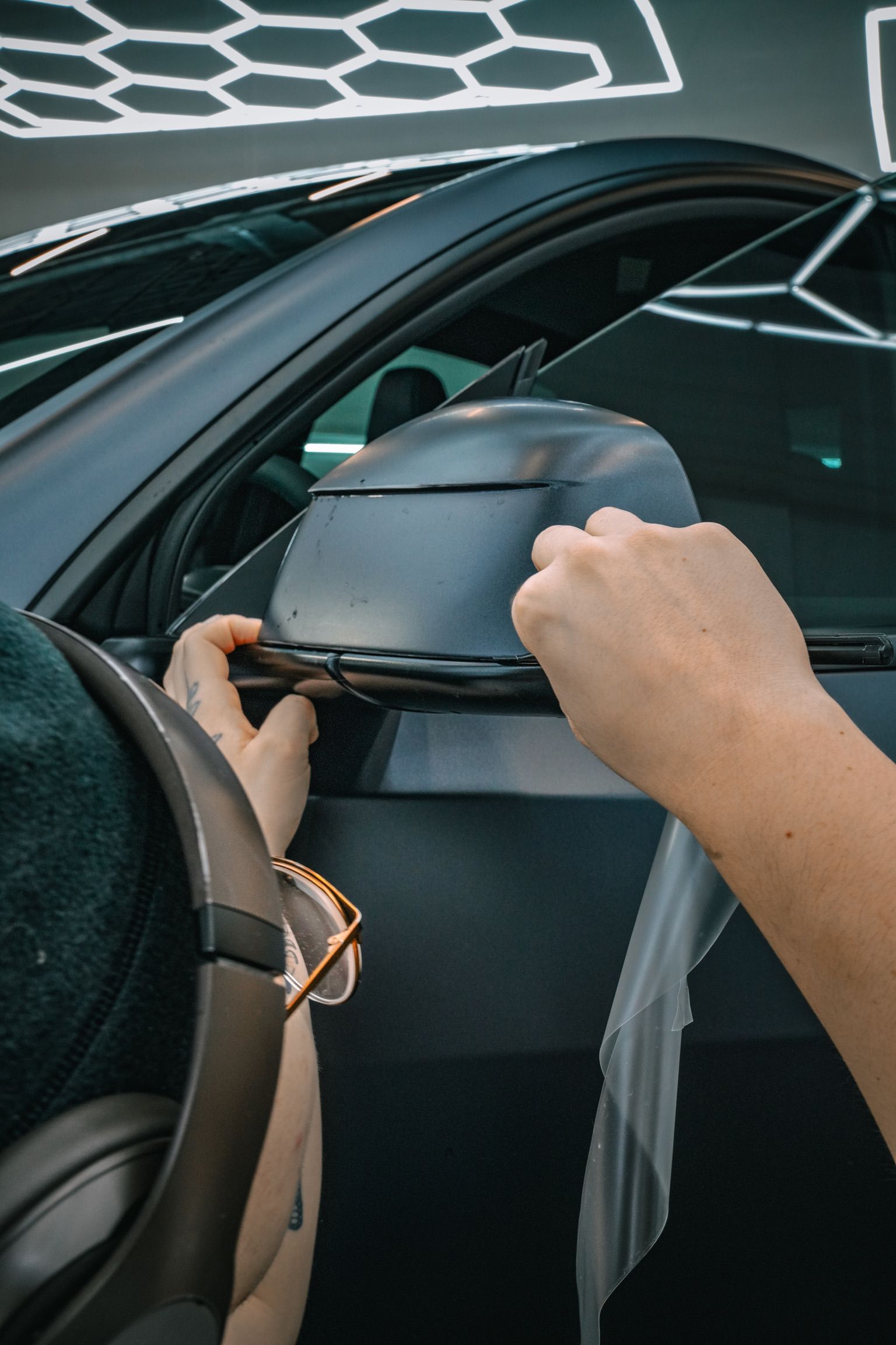 Hands applying a clear protective film to the side mirror of a matte black car.