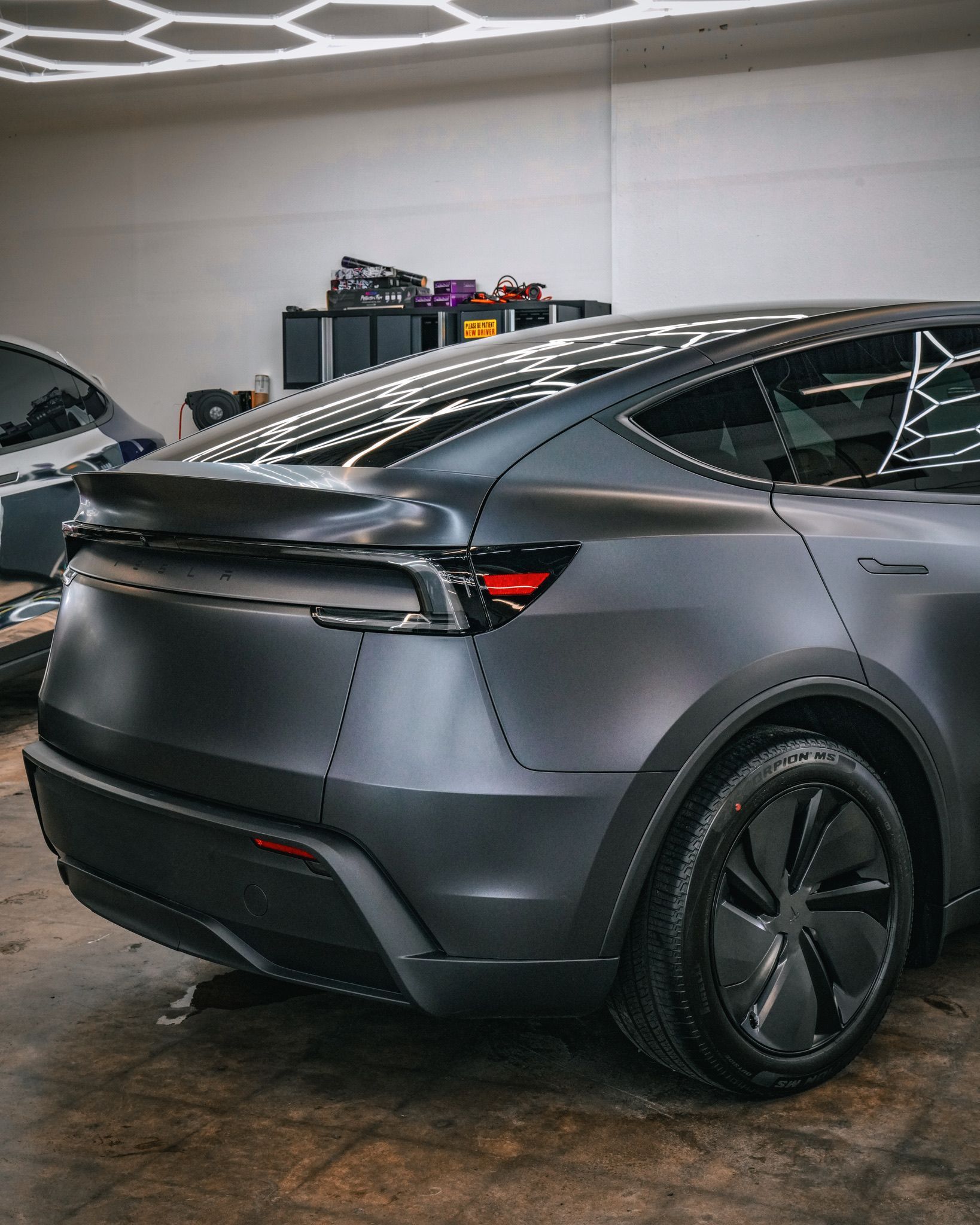 Rear angle view of a matte charcoal Tesla Model Y with a customized, industrial-style riveted wrap in a bright garage.