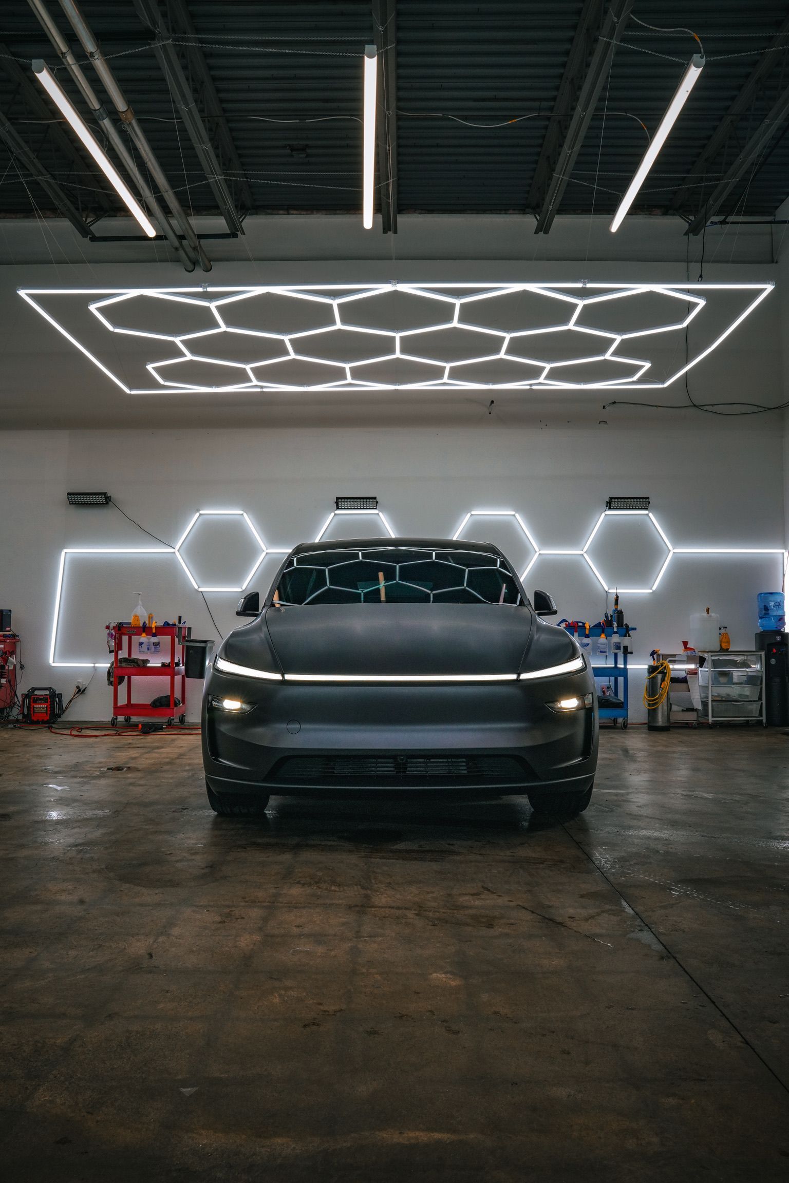 A matte dark car parked in a garage with a neon hexagonal ceiling light grid and geometric wall lighting.