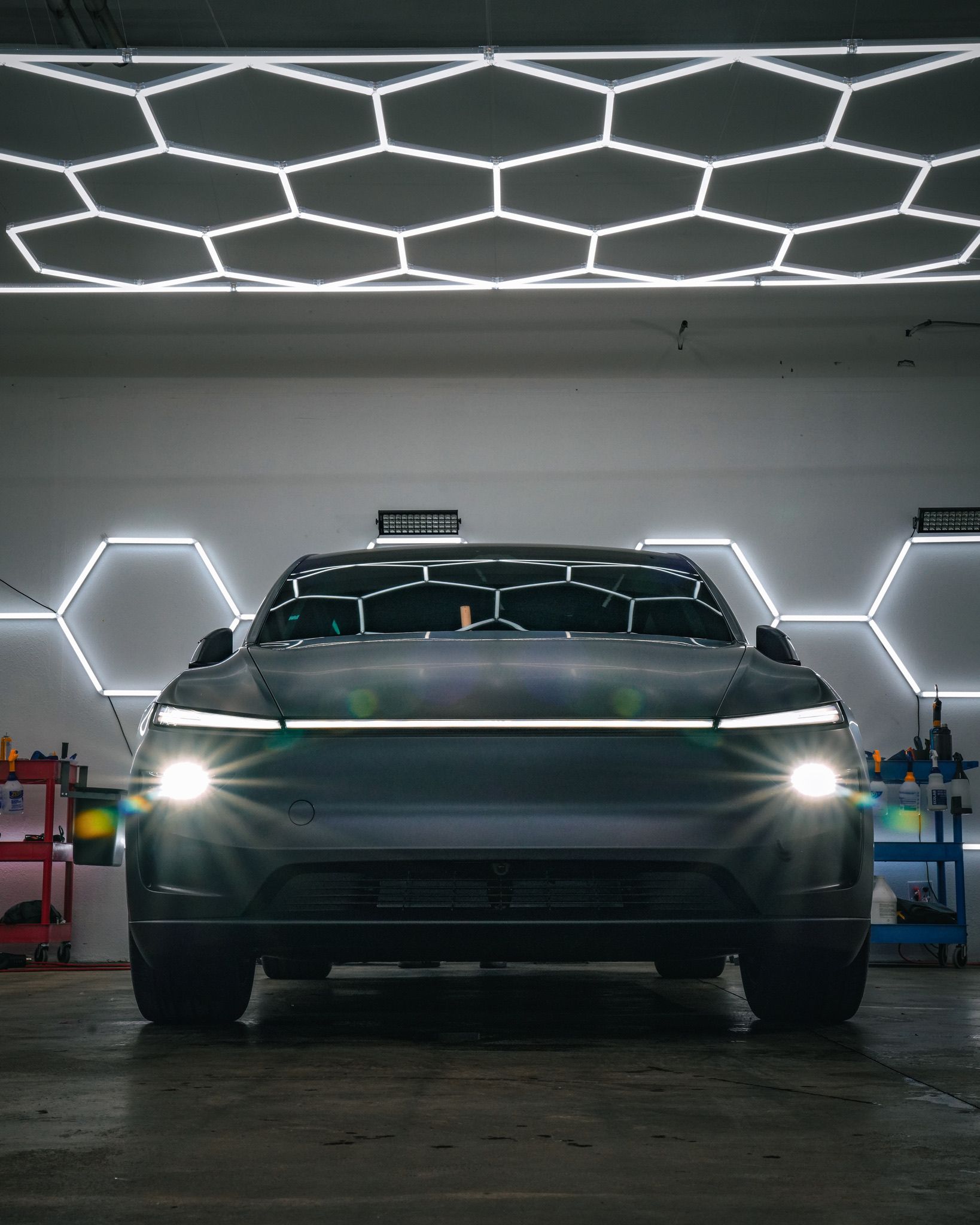 A grey car sits in a garage with illuminated, hexagonal honeycomb-patterned lights on the ceiling and walls.