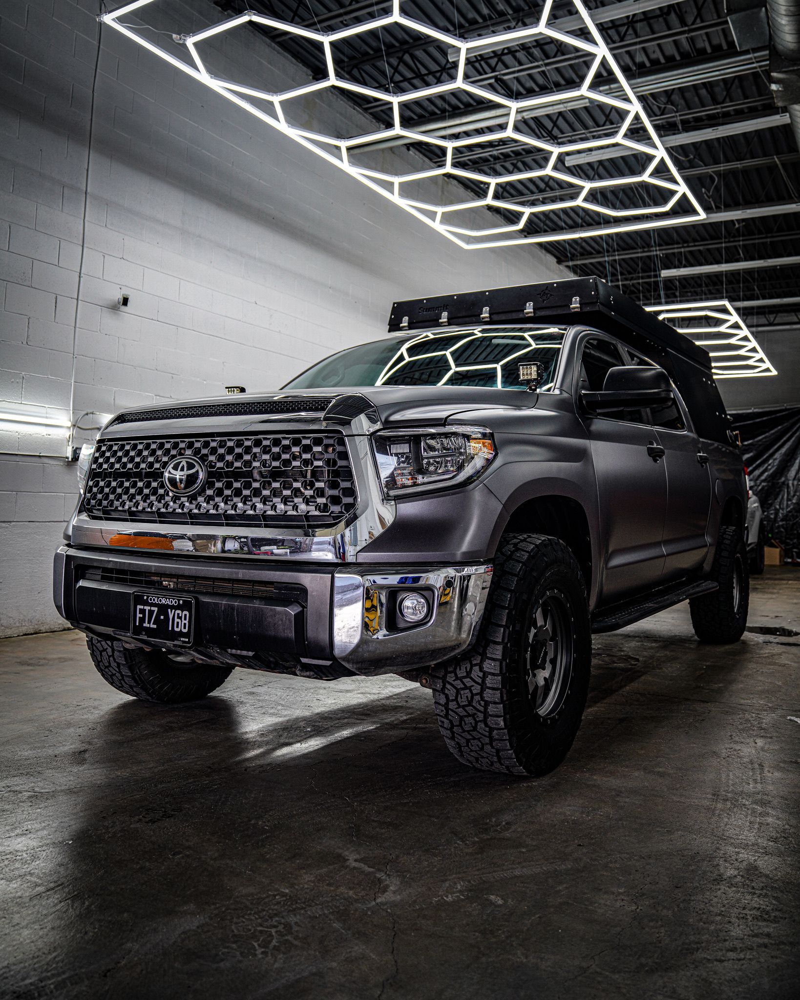 A matte gray Toyota Tundra pickup truck with off-road tires and a roof rack parked in a garage with hexagonal ceiling lights.