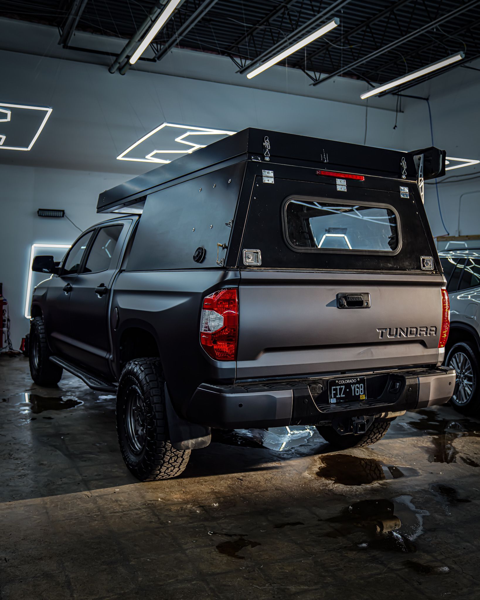 Matte black Toyota Tundra pickup truck with a custom, riveted camper shell parked in a garage with overhead neon lights.