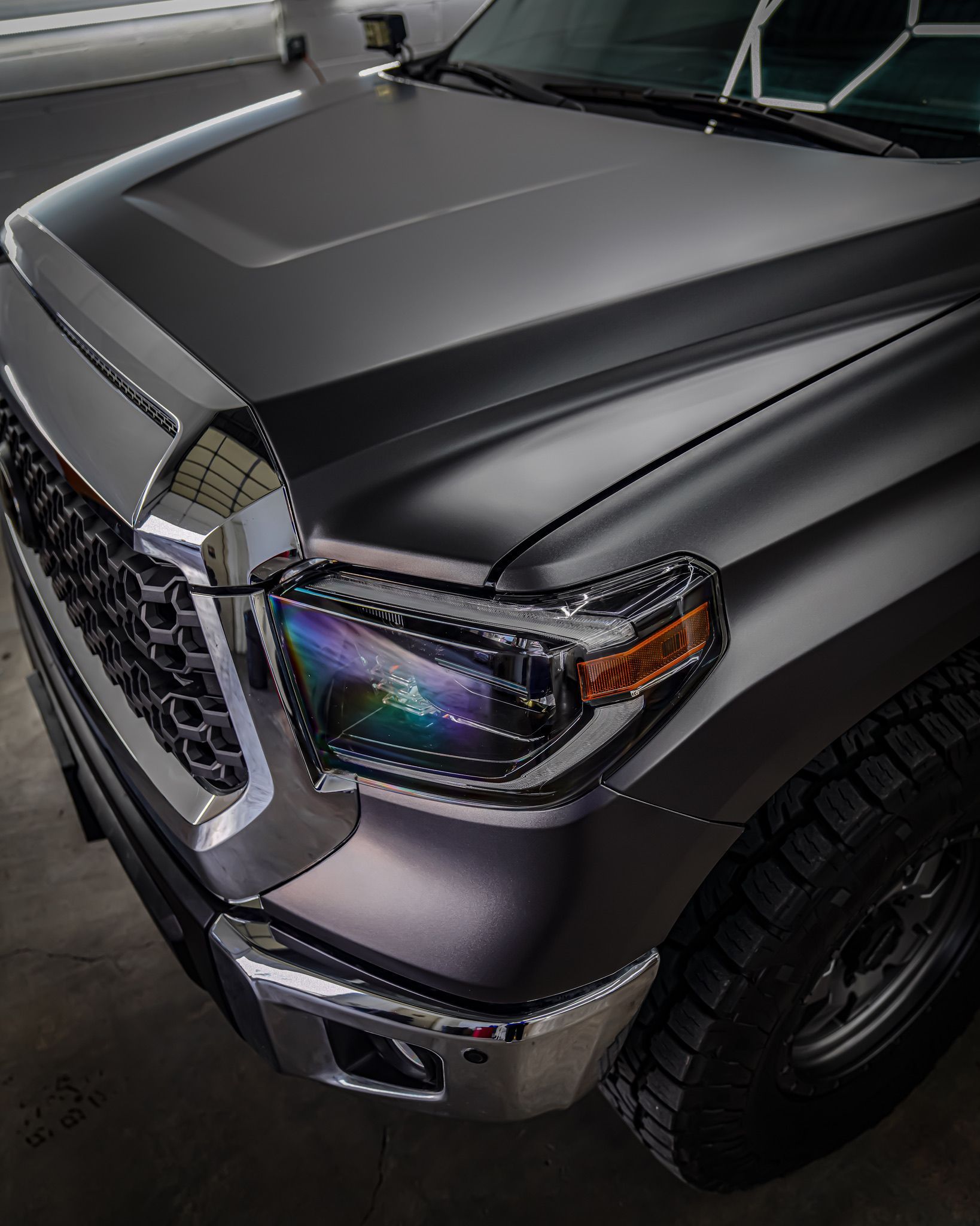 A high-angle view of a matte grey Toyota Tundra truck's front end, showing the grille, headlight, and hood.