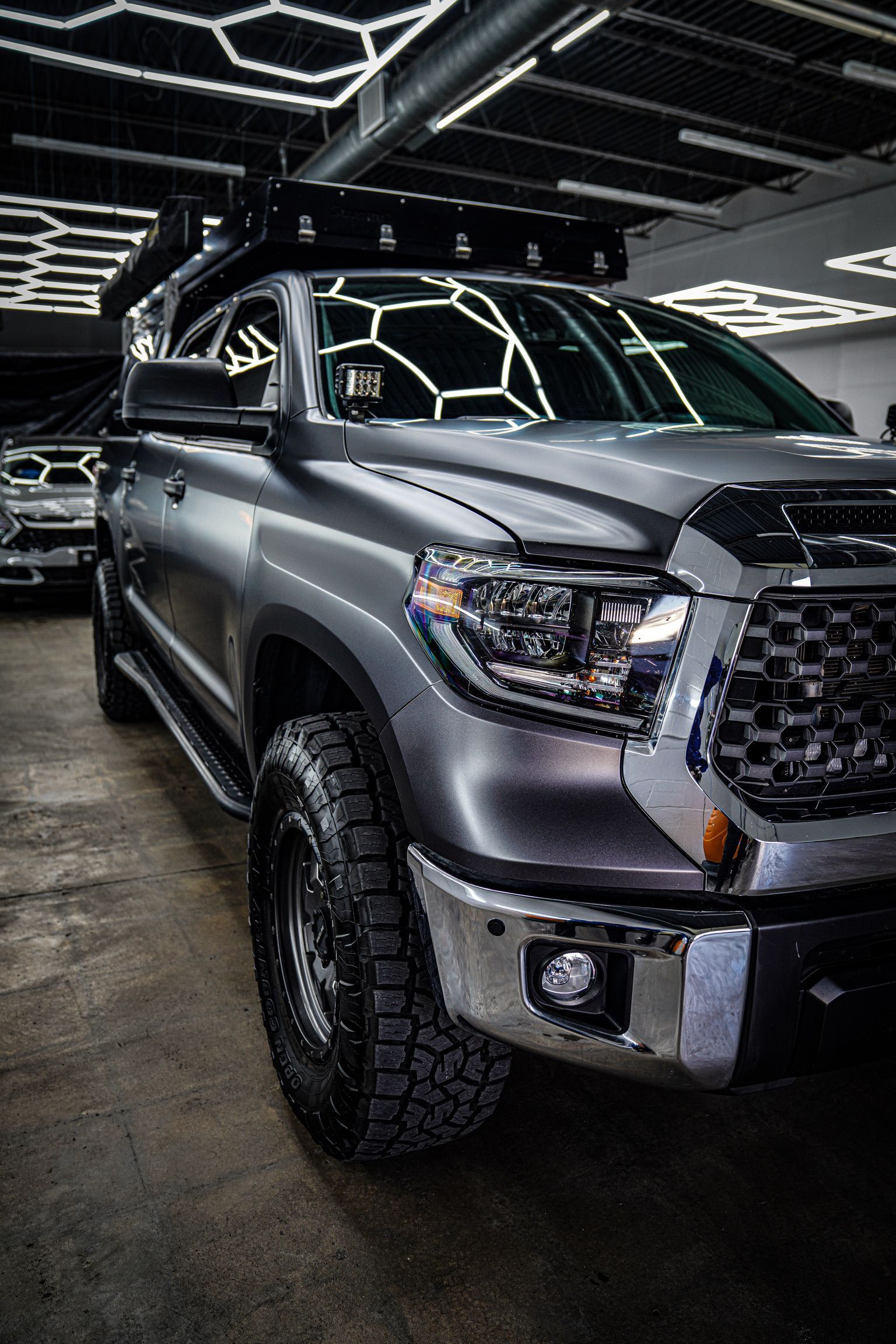 A matte grey Toyota Tundra with an off-road roof rack and rugged tires parked in a garage with geometric ceiling lights.