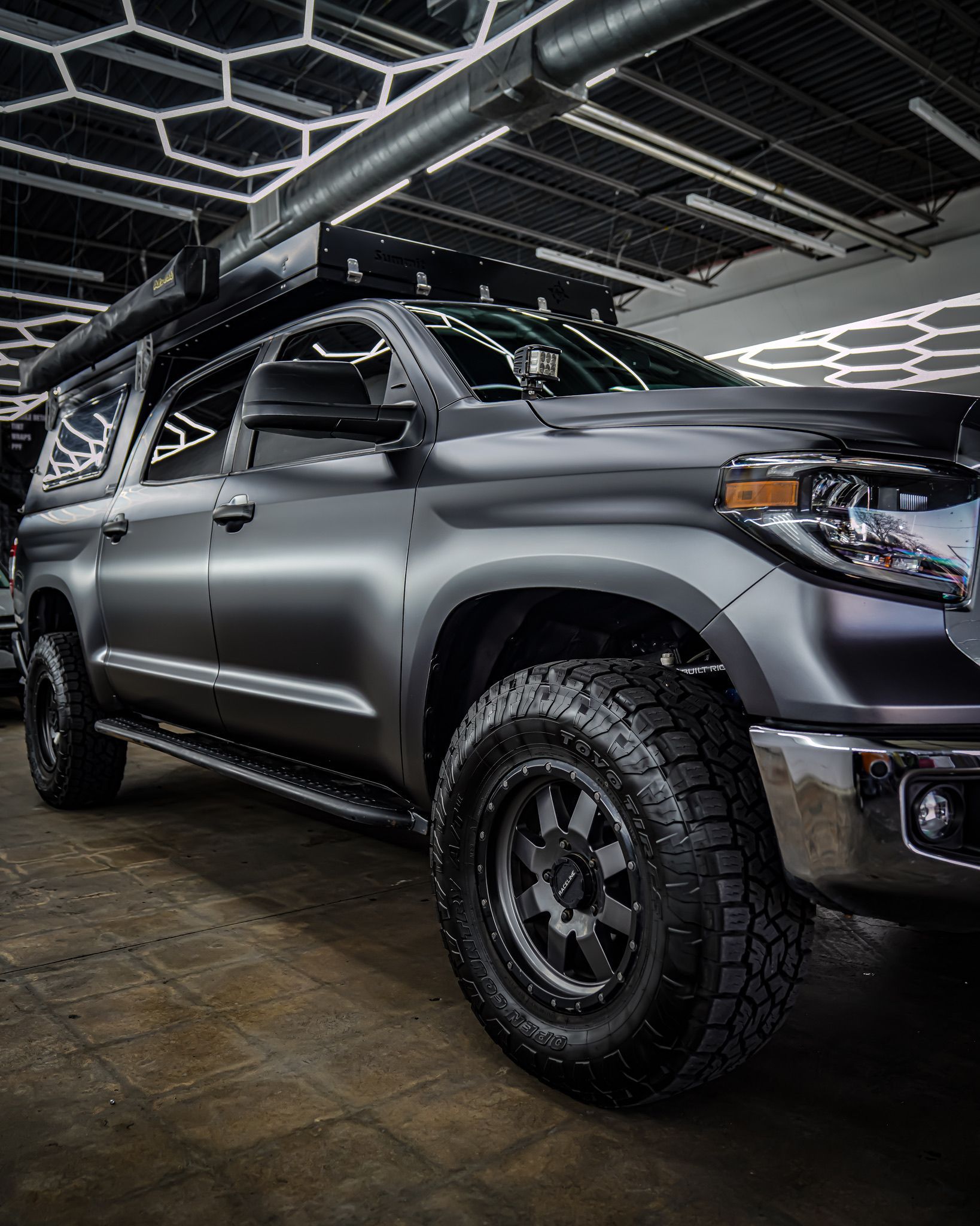 A matte black Toyota Tundra pickup truck with off-road tires and a roof rack parked in a garage with geometric lighting.