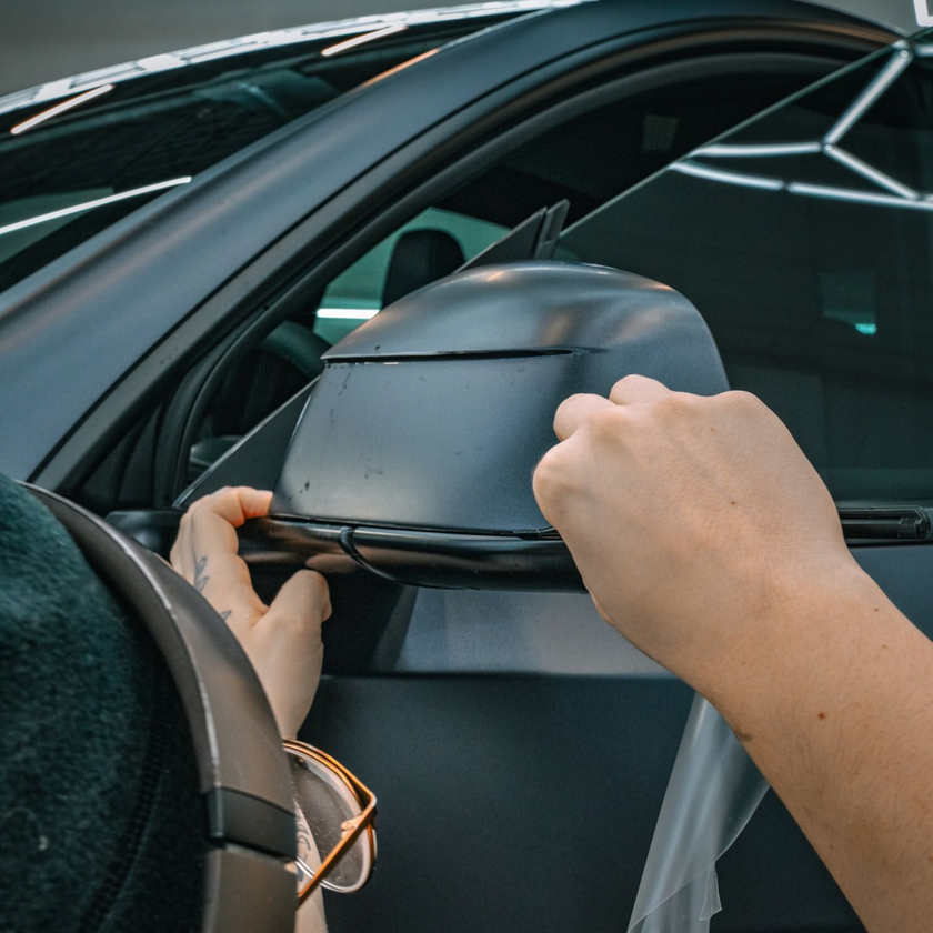 Two hands carefully applying a protective vinyl wrap onto the side mirror of a matte black car.