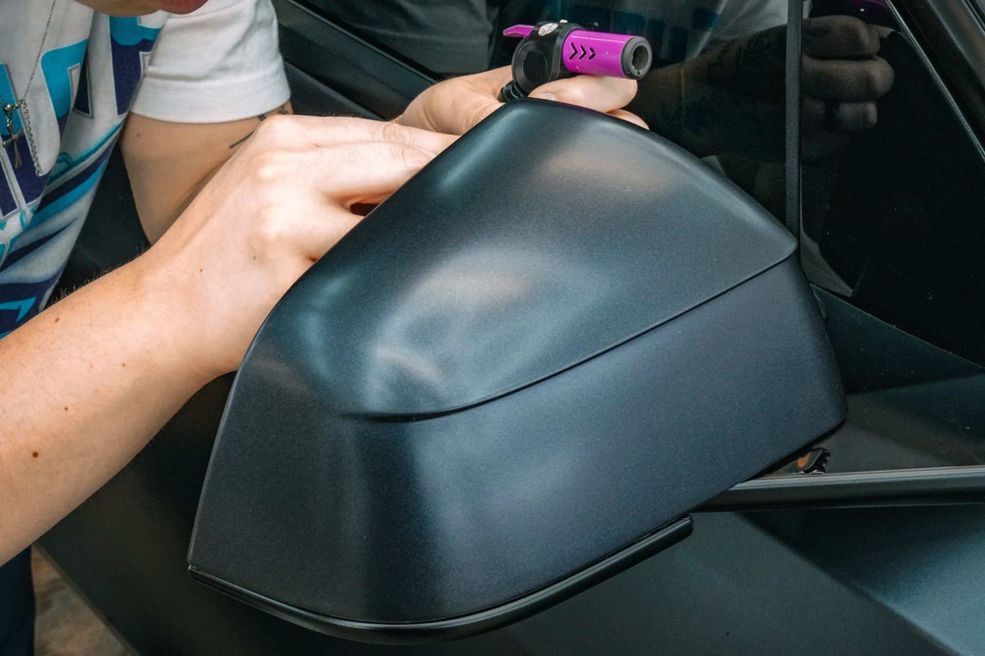 Technicians apply a clear protective film onto the hood of a black car in a garage with specialized overhead lighting.