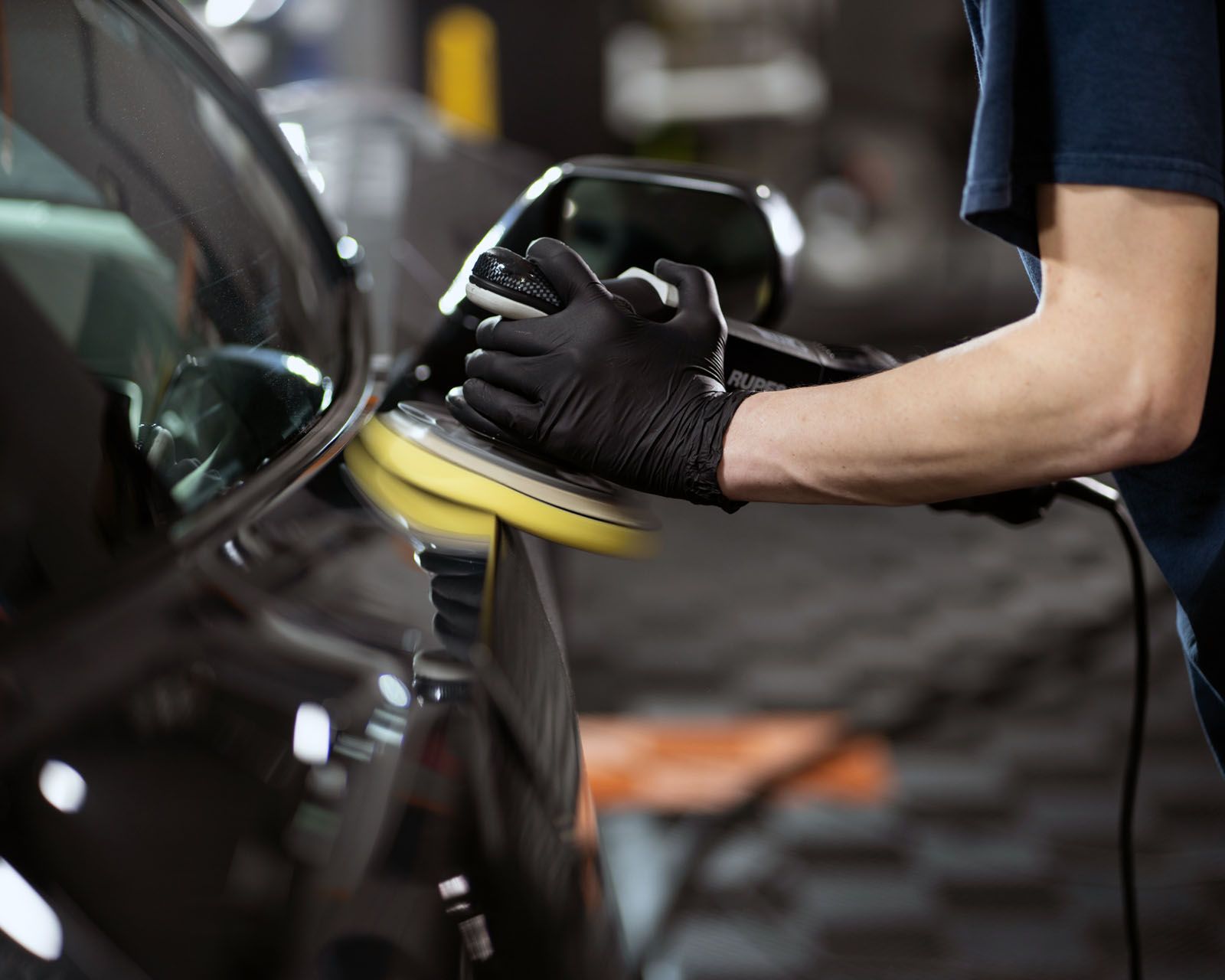A person wearing black gloves uses a power buffer with a yellow pad to polish the side of a black car in a workshop.
