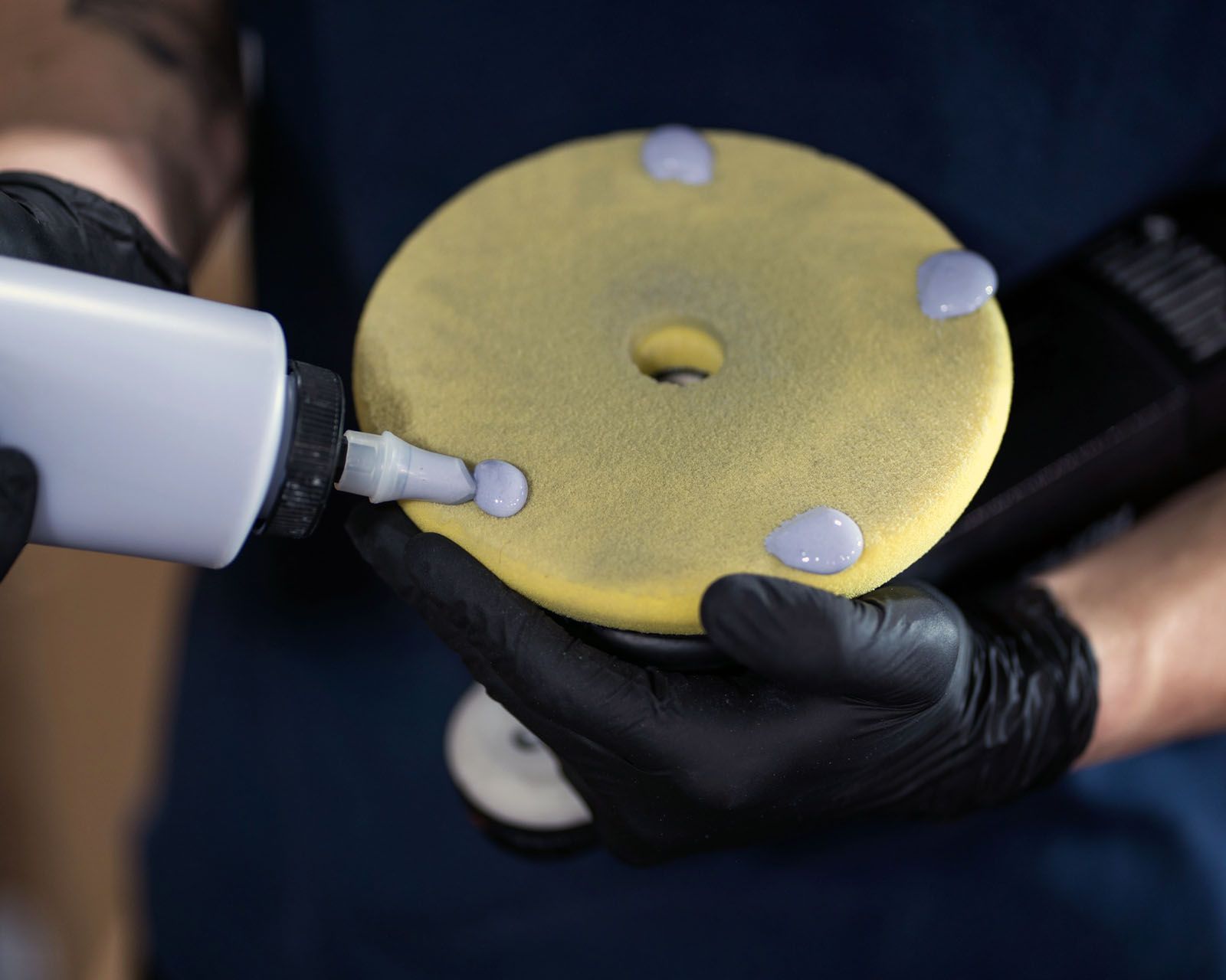A person wearing black gloves applies drops of gray polishing compound to a yellow foam buffing pad.