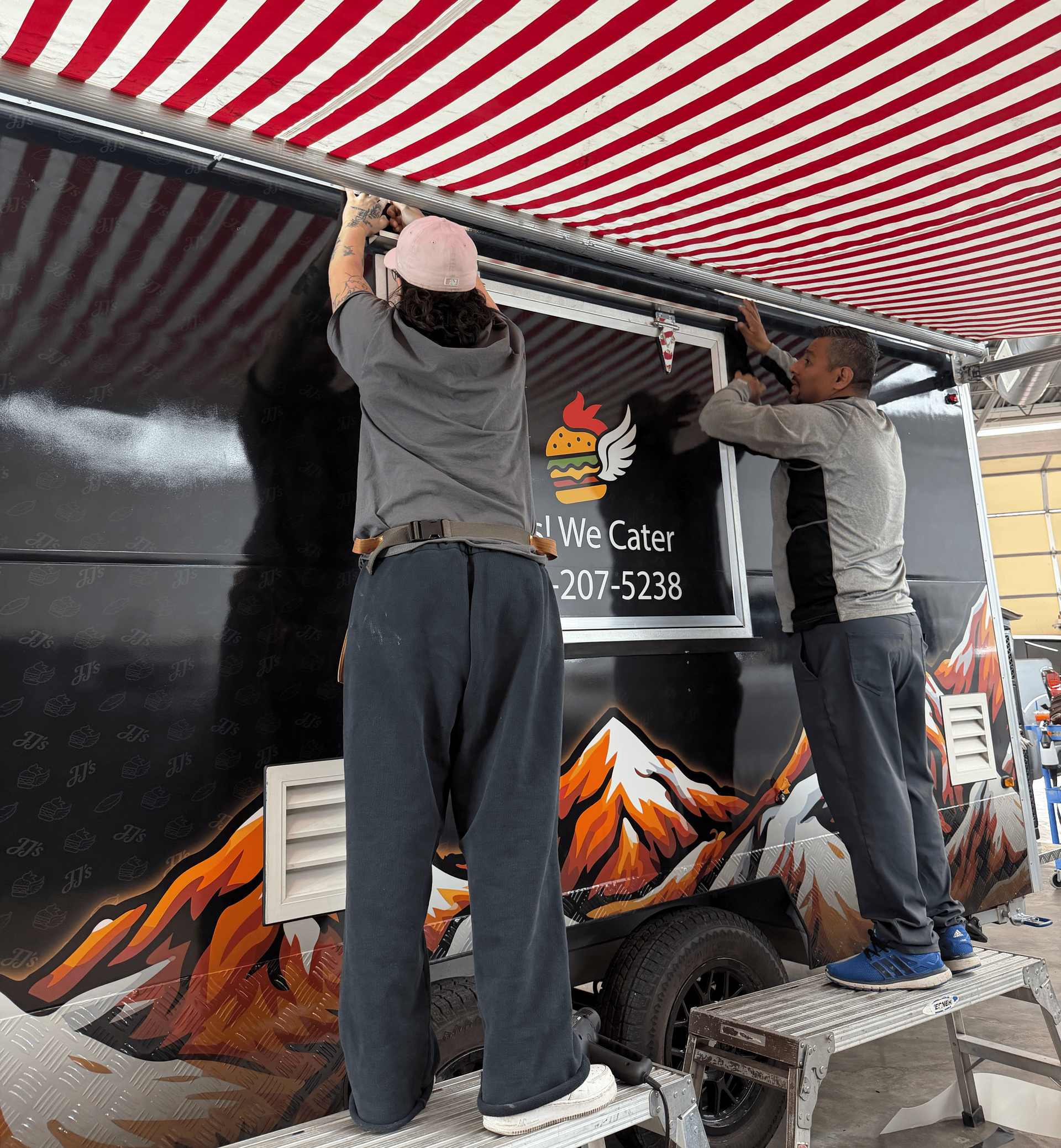 Two people install a red-and-white striped awning on a food truck with mountain graphics.