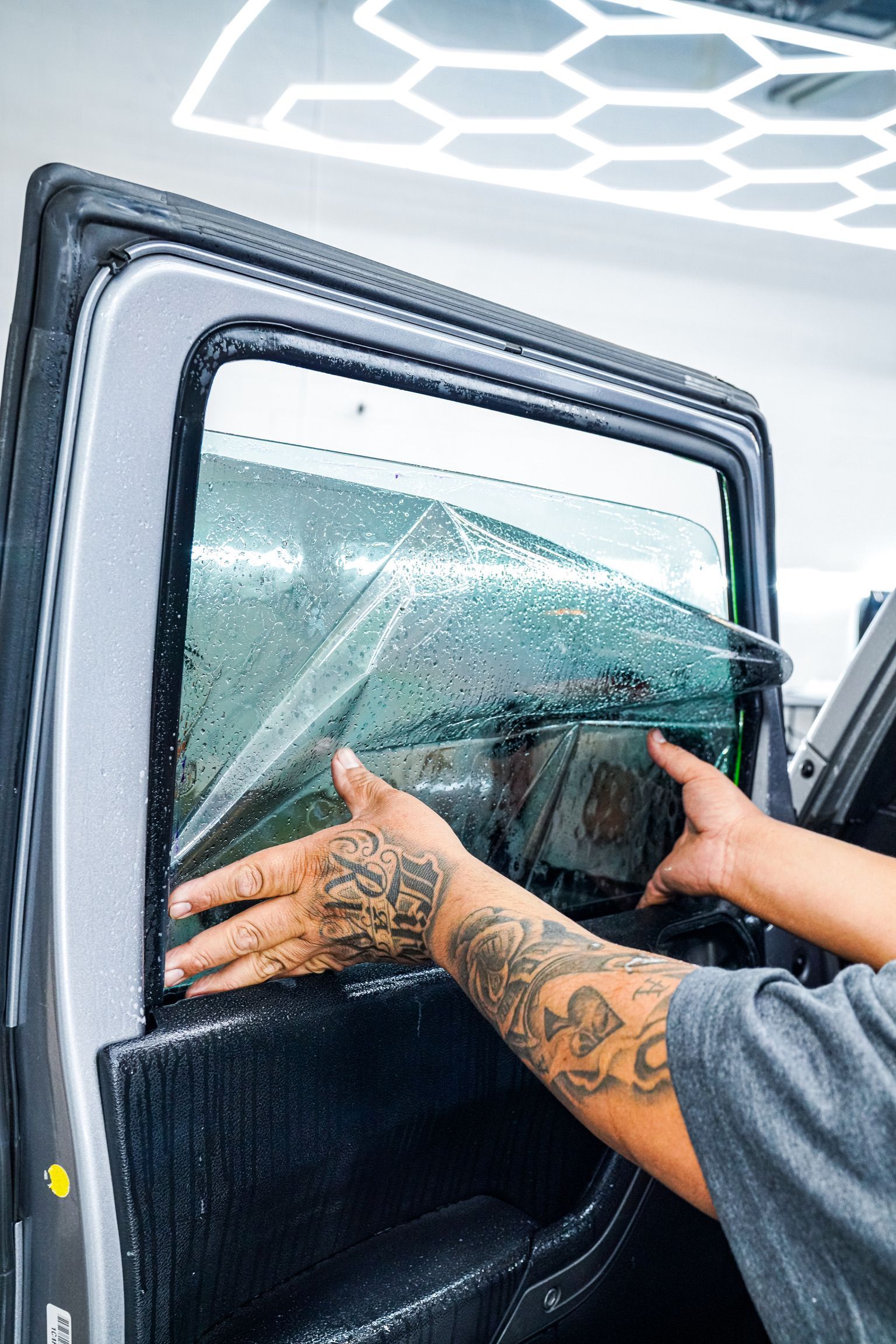 A tattooed technician carefully applies a clear window tint film onto a car door window in a well-lit workshop.
