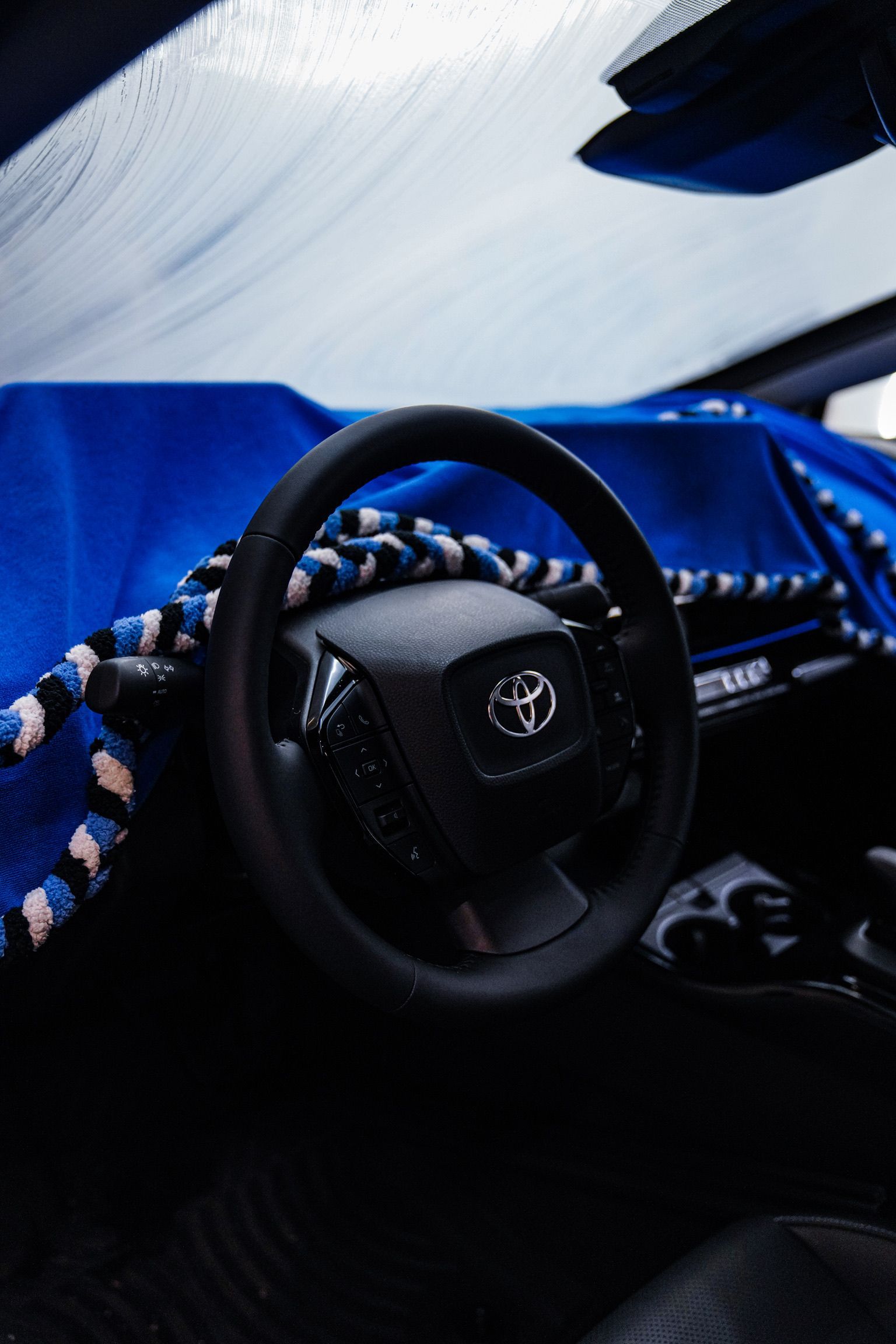A person kneels to apply a protective film to the front bumper of a blue sports car in an auto shop.