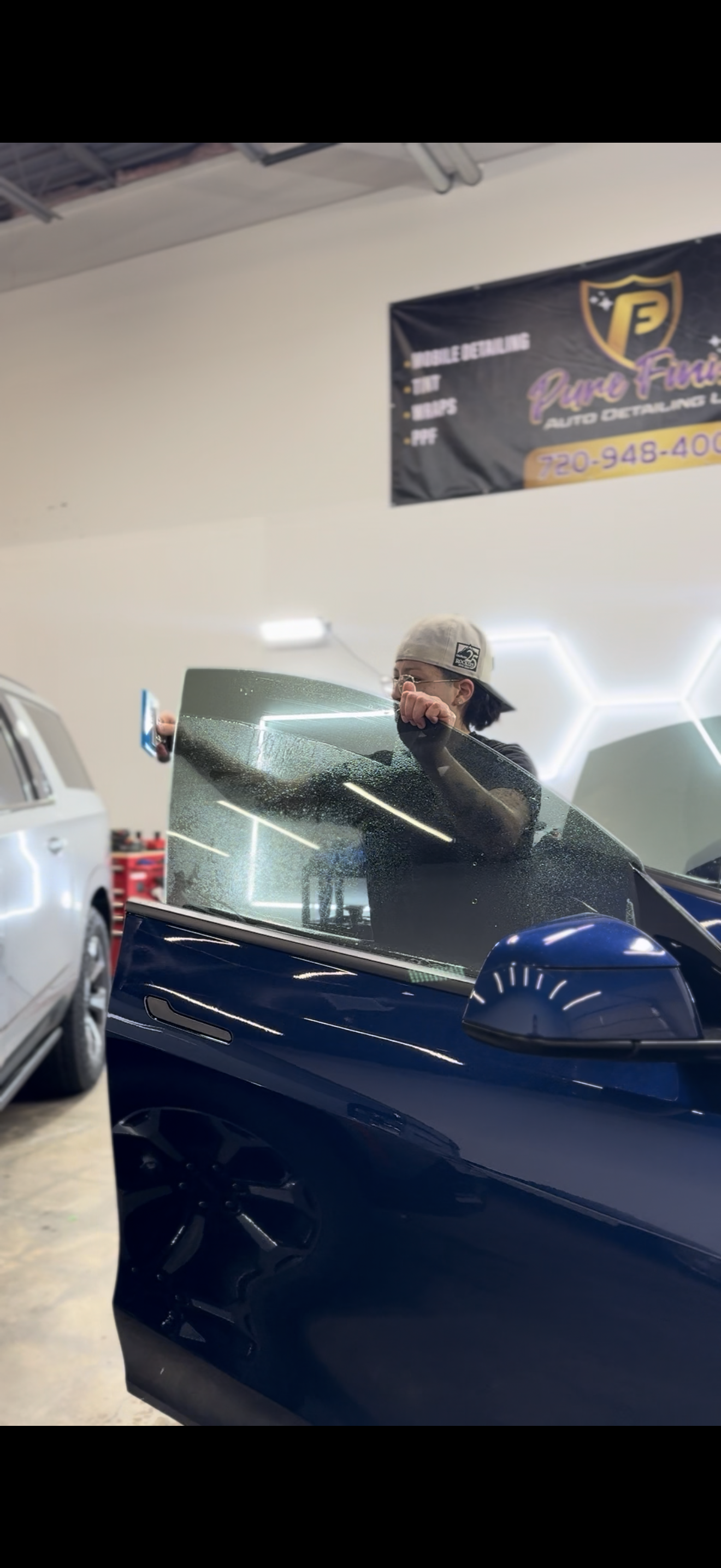 A person in a shop wearing a cap installs a dark window tint film onto the front door window of a navy blue vehicle.