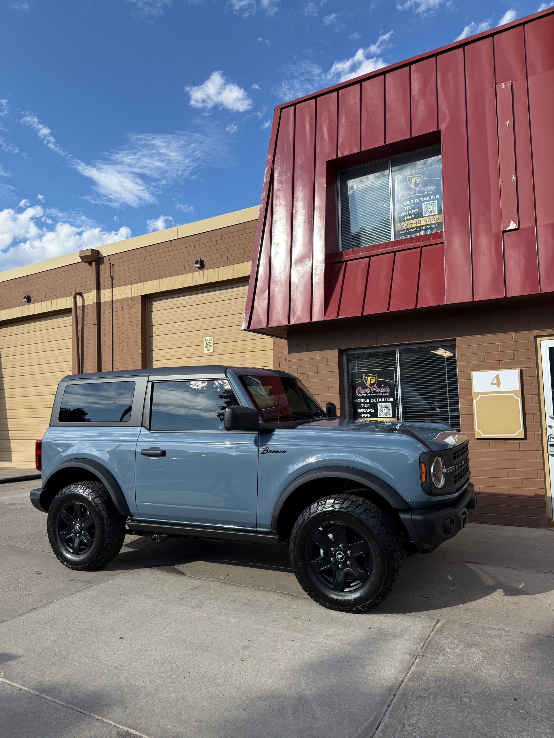 A two-door gray Ford Bronco parked on an asphalt lot in front of a brick commercial building with a red metal roof.