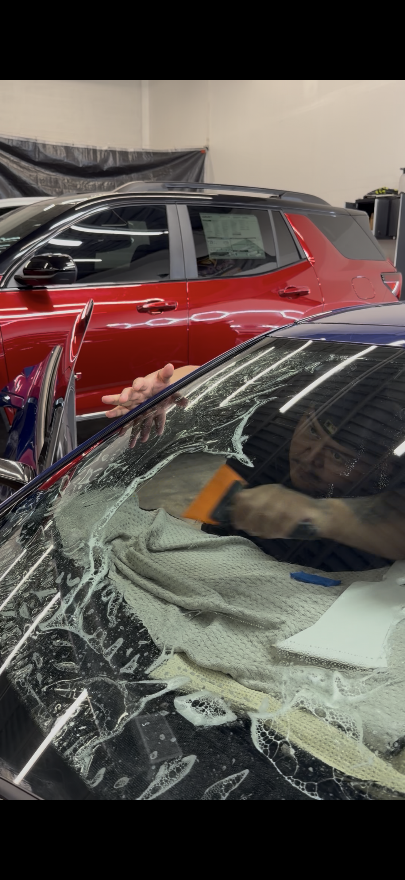 A technician in a garage uses a squeegee to apply film to a car windshield, with a red SUV parked behind them.