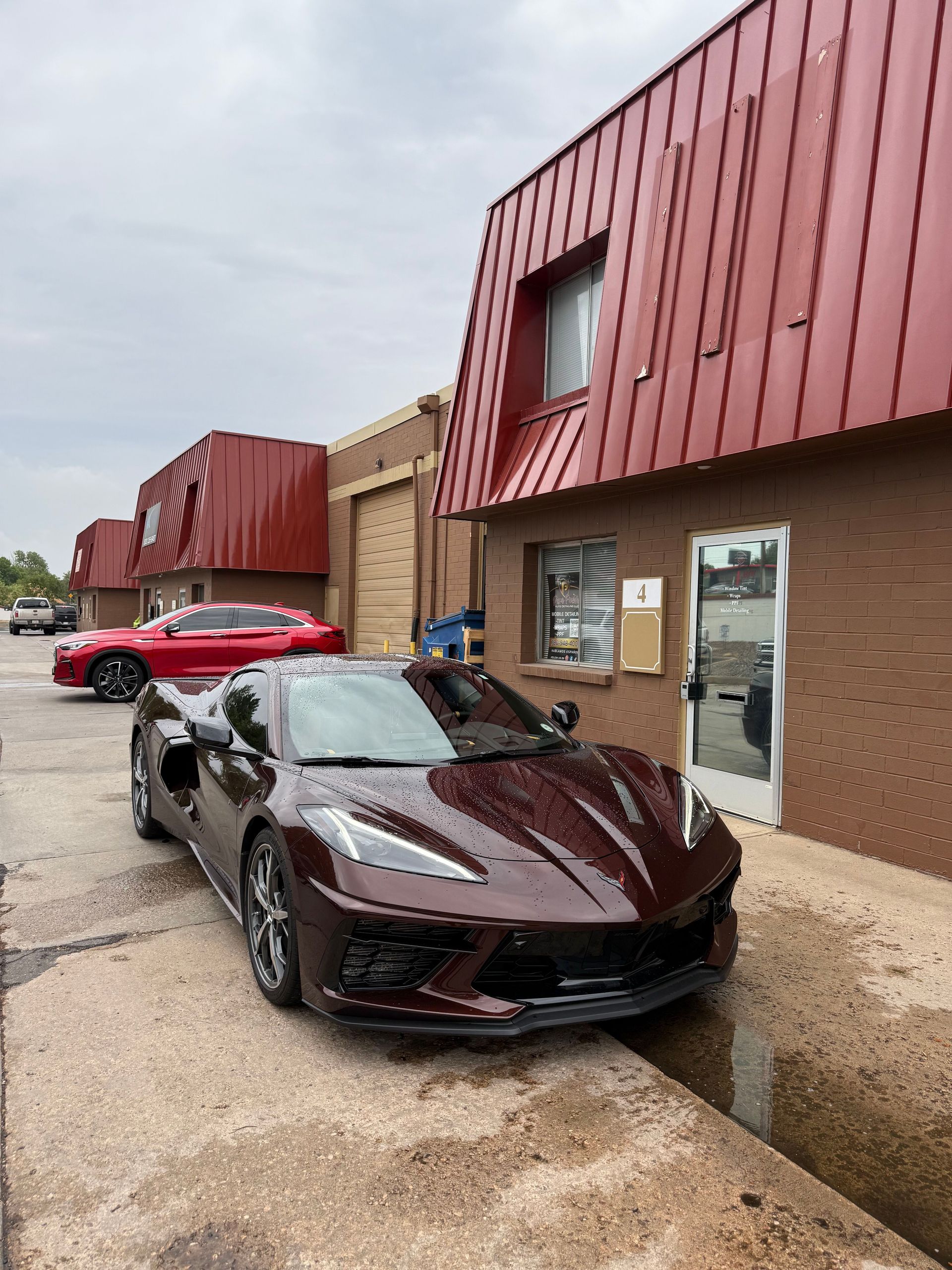 A dark red Corvette is parked on concrete next to a brick building with a red metal roof. Another red car is in back.